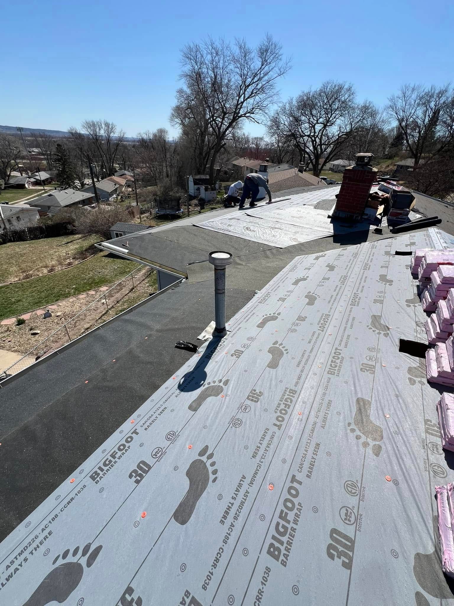 Roof under construction with gray underlayment, vents, and workers near a residential neighborhood