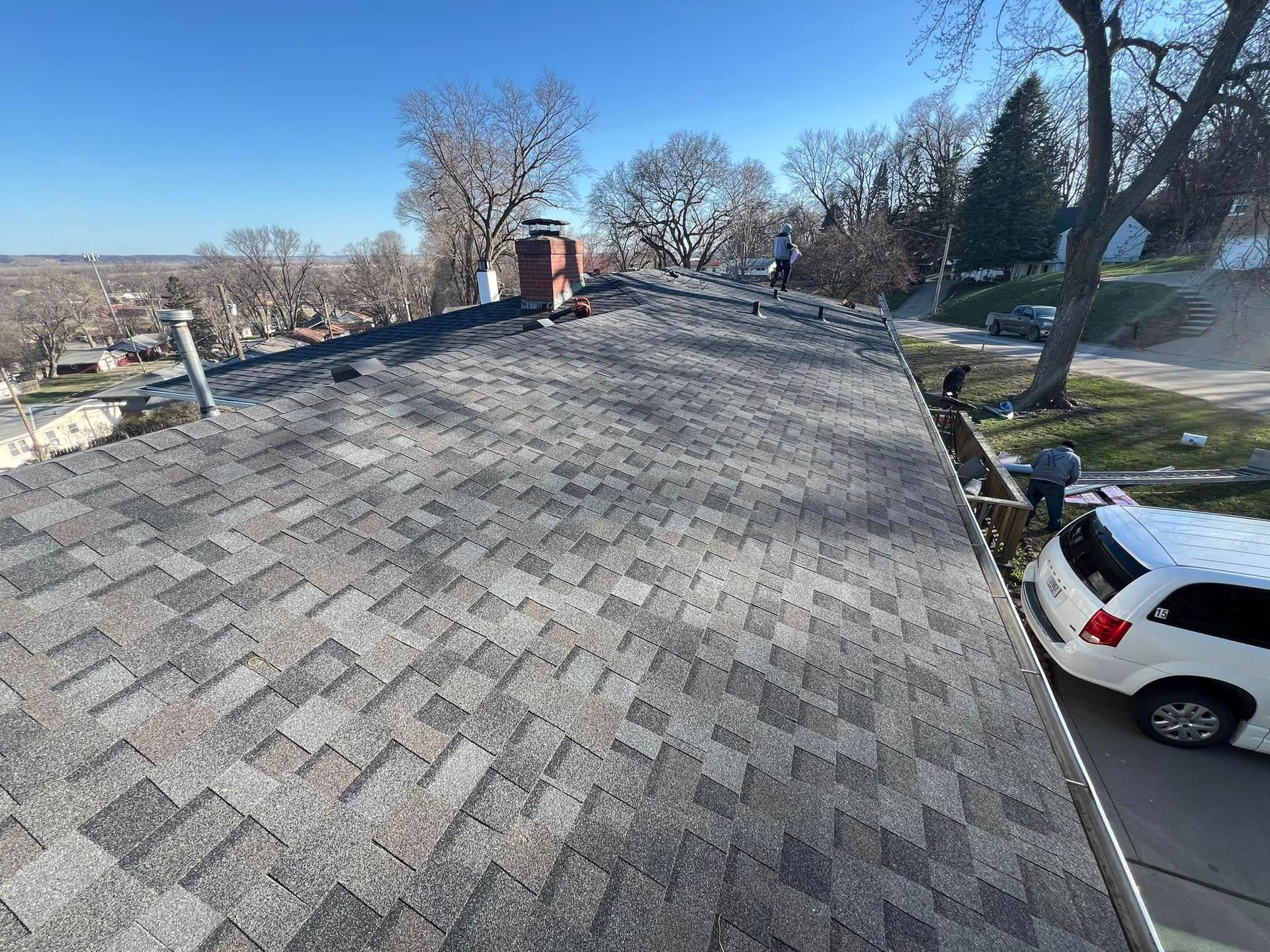 Rooftop covered in gray shingles beside a street, with trees, houses, and a parked white car below.