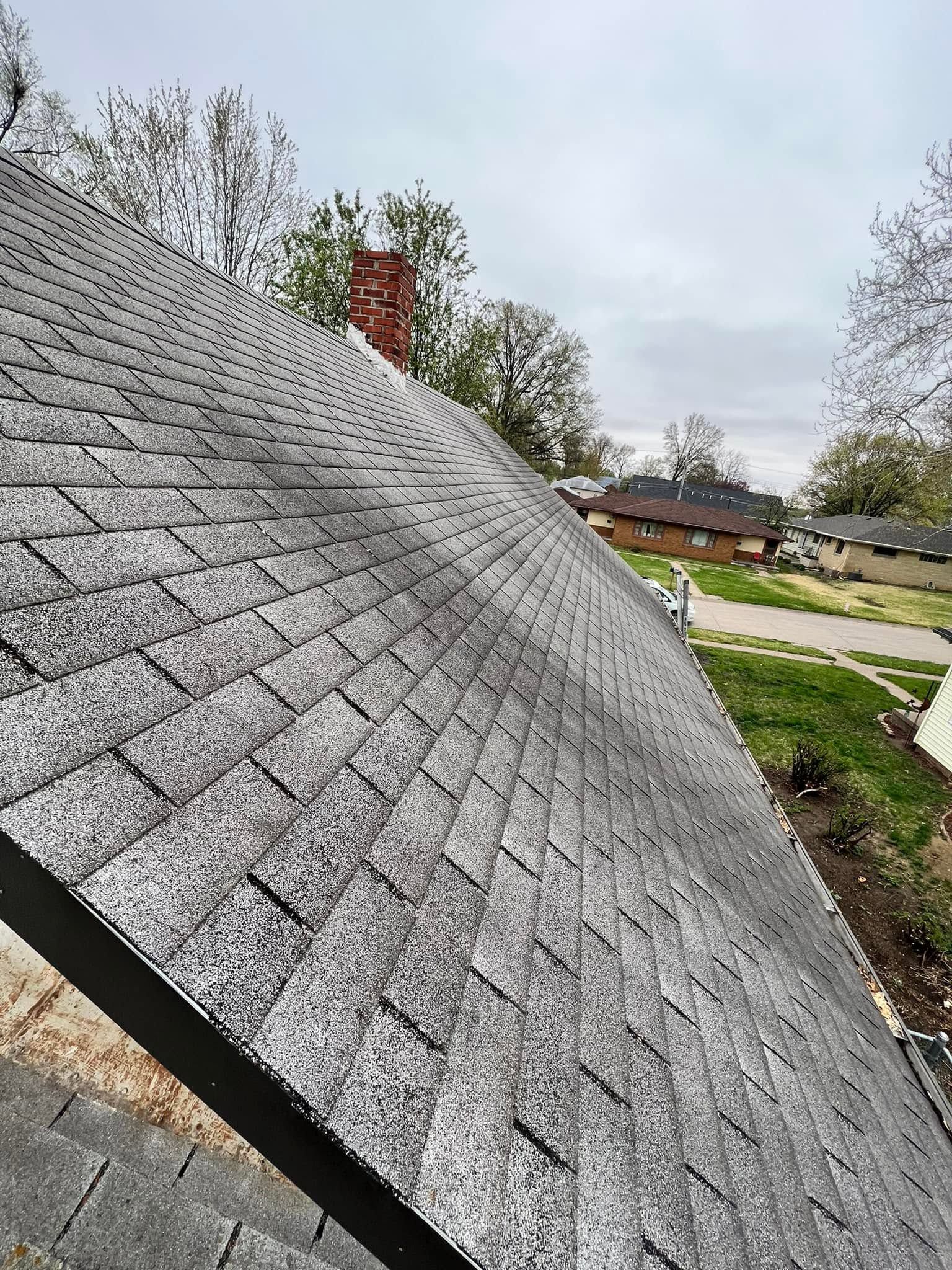 Close-up of a gray shingled roof beside a grassy yard, with trees and houses in the background.