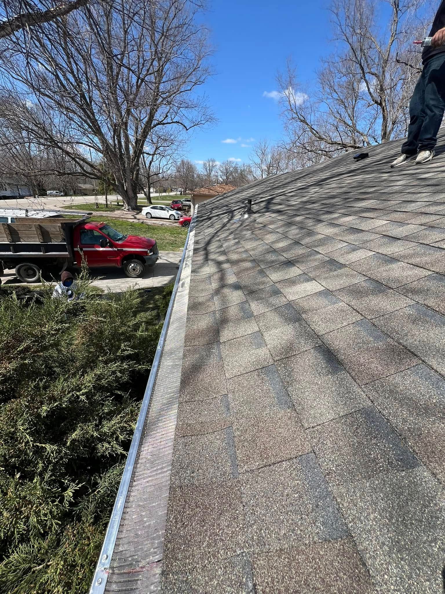 Person on a steep shingled roof beside a truck and leafless tree on a sunny day