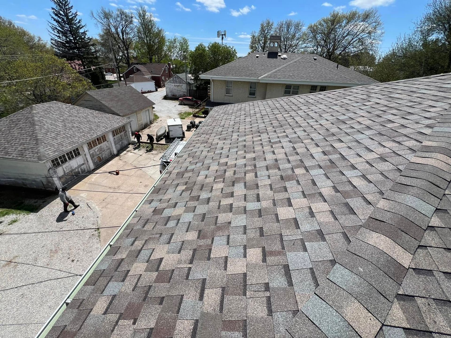 View across gray shingled rooftops of a house under roof repair, with a yard and trees beyond.