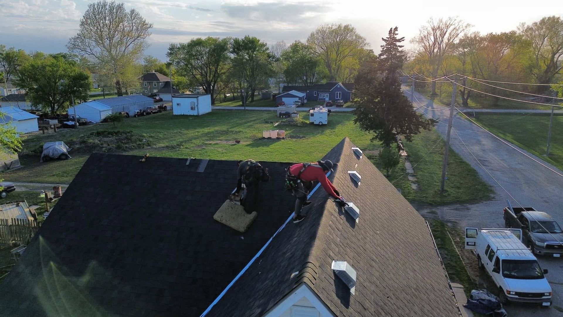 Worker on a roof repairing shingles beside a ladder in a suburban neighborhood at sunset