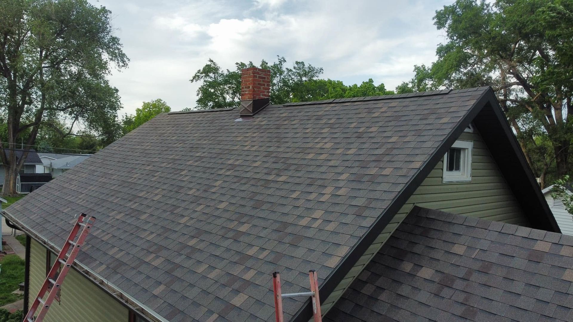 Roofing shingles being installed on a steep house roof with ladders and a brick chimney.