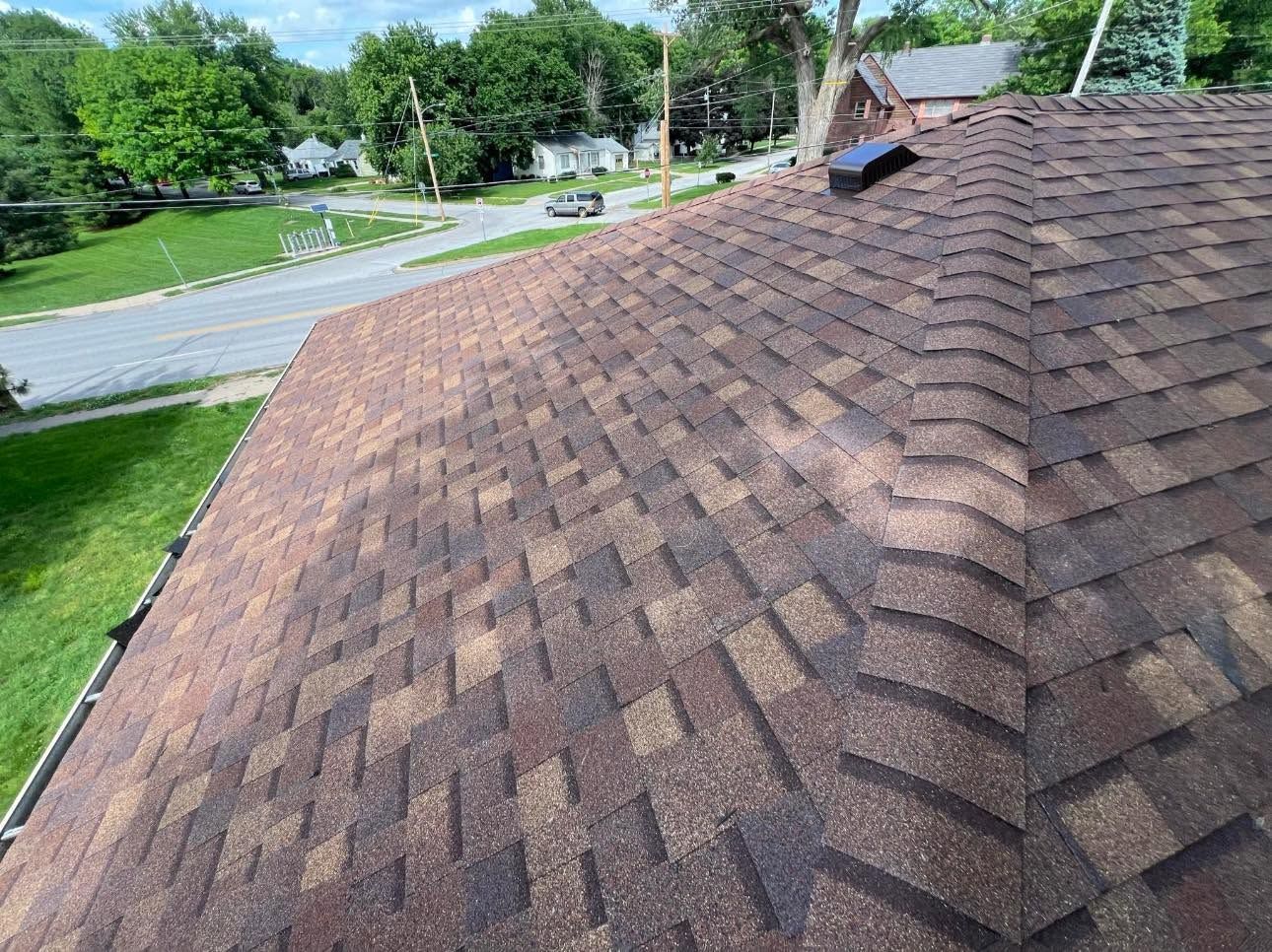 Rooftop with brown shingles beside a leafy suburban street under a clear sky