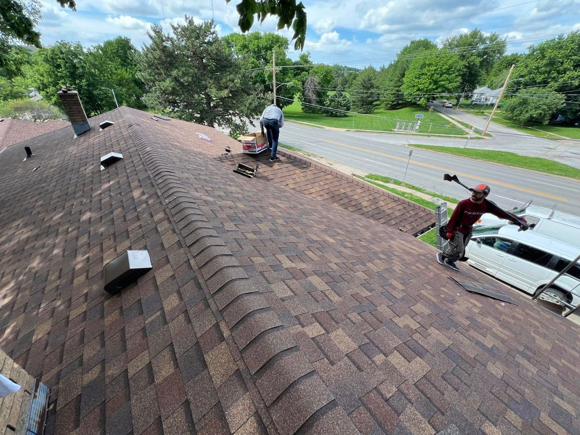 Workers repairing a shingled roof beside a tree-lined road on a sunny day