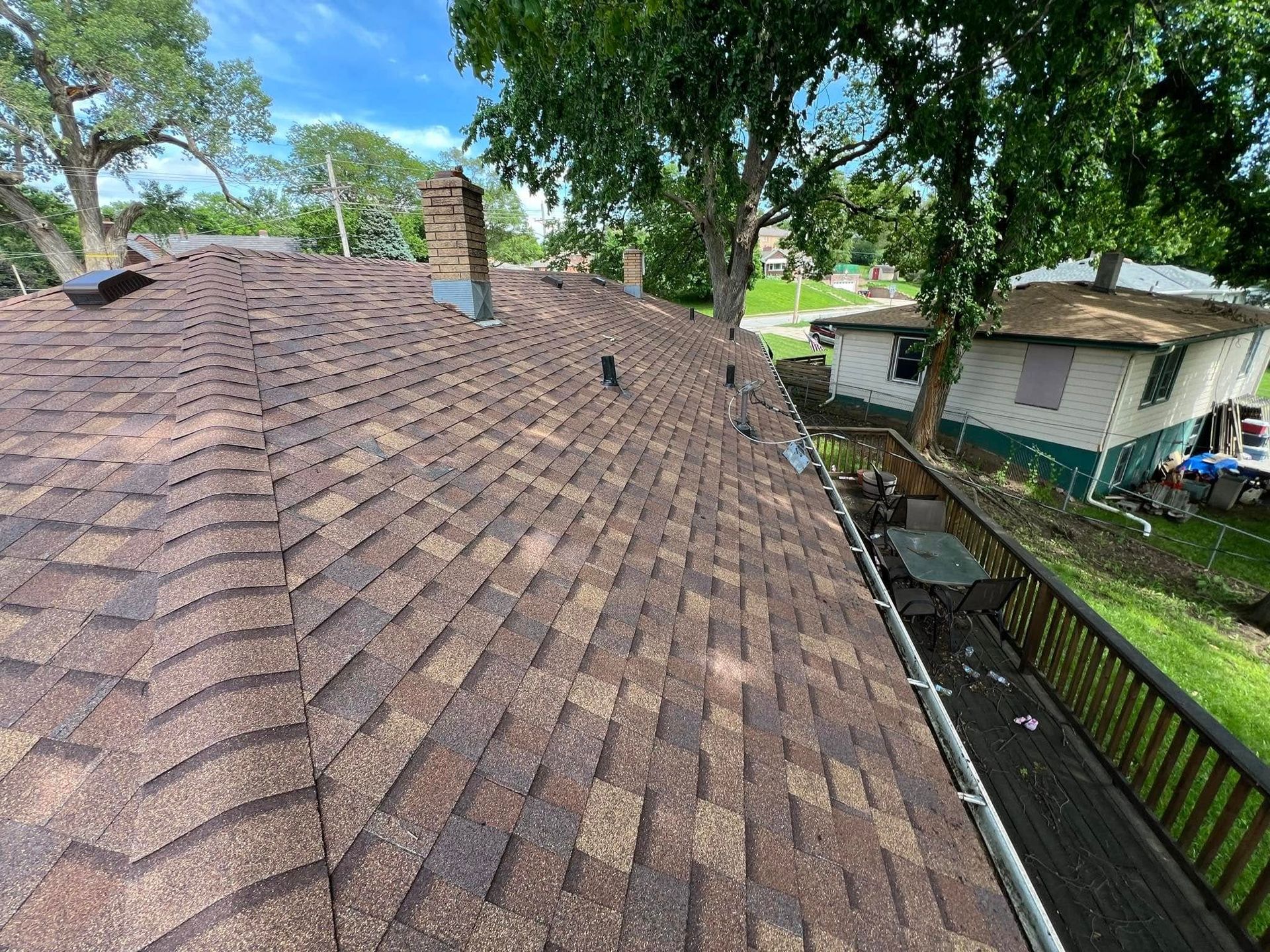 Brown shingled roof overlooking a yard, trees, and a house on a sunny day