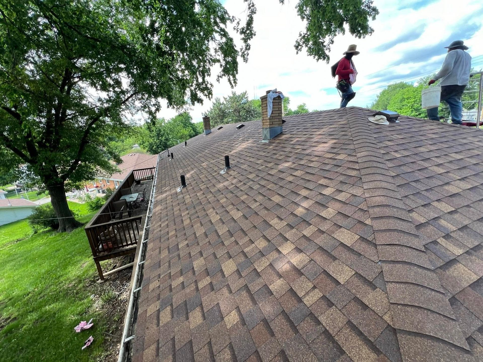 Workers on a shingled roof, with one standing near a chimney on a tree-lined residential property.