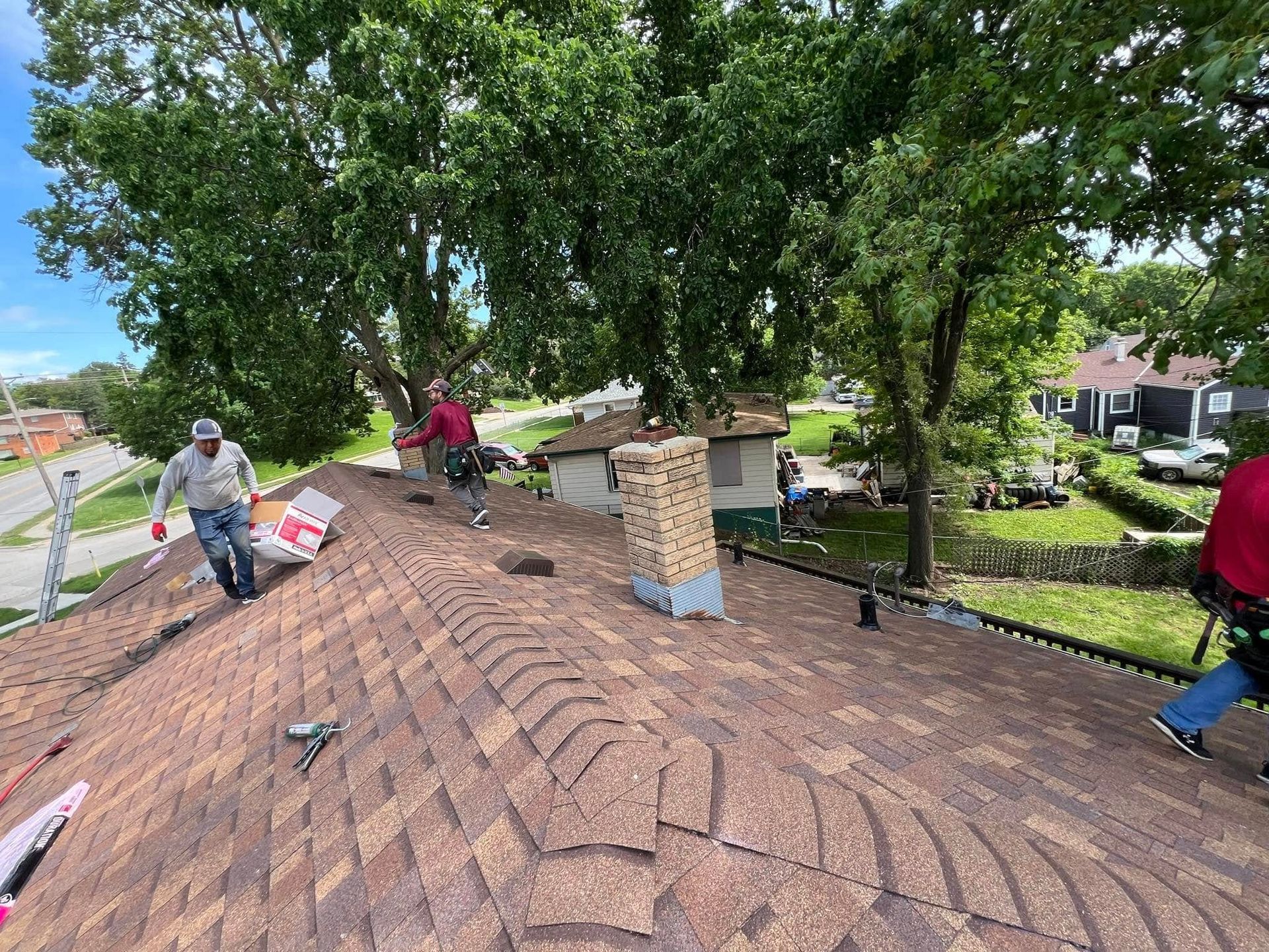 Workers installing shingles on a sloped roof beneath large trees, with a brick chimney in the center