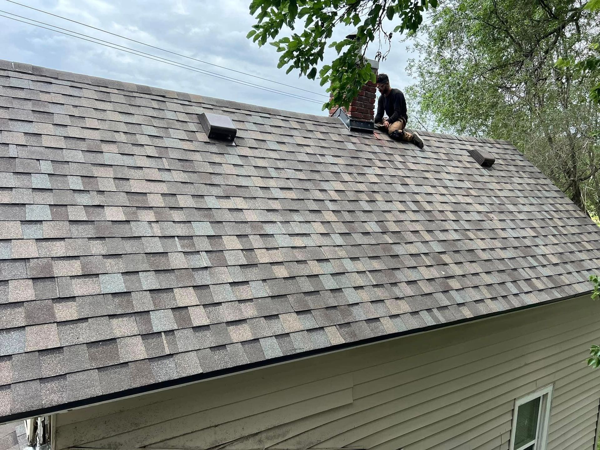 Two workers repairing a shingled roof beside a tree-lined house.