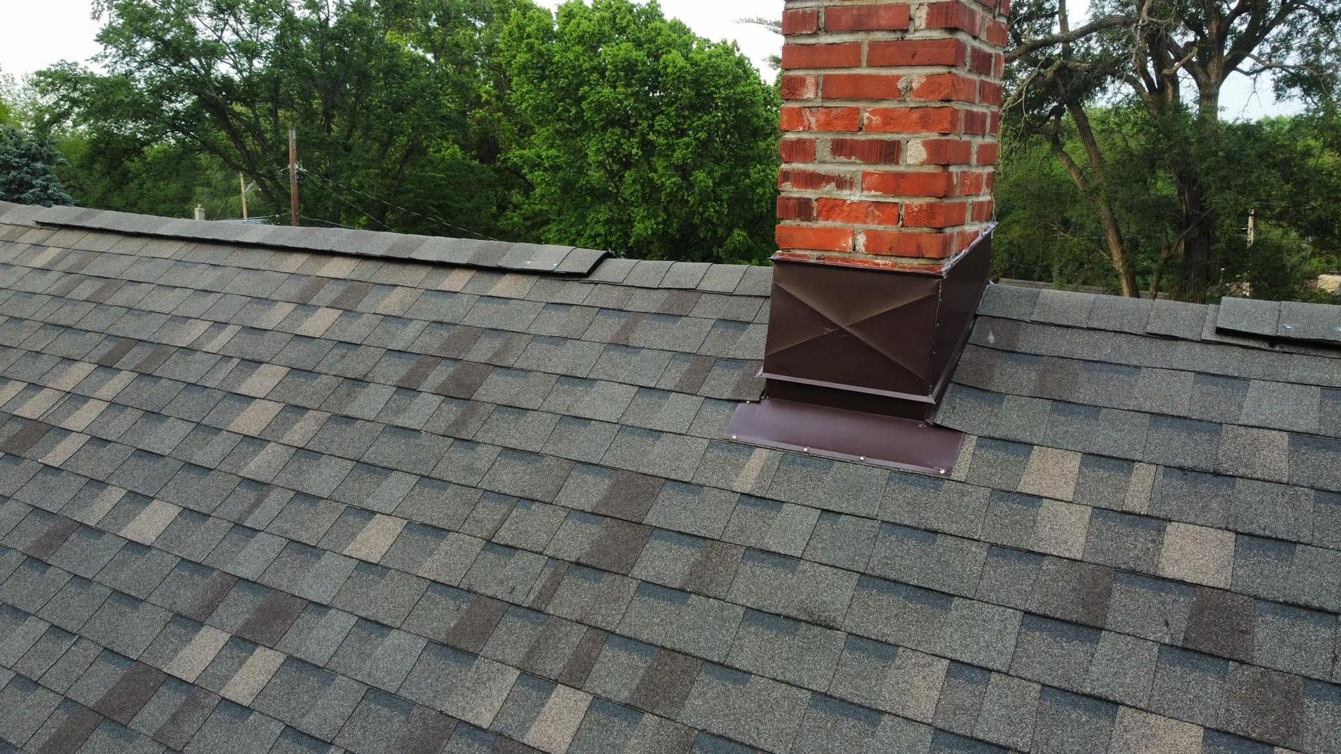 Shingled roof with brick chimney flashing and green trees in the background