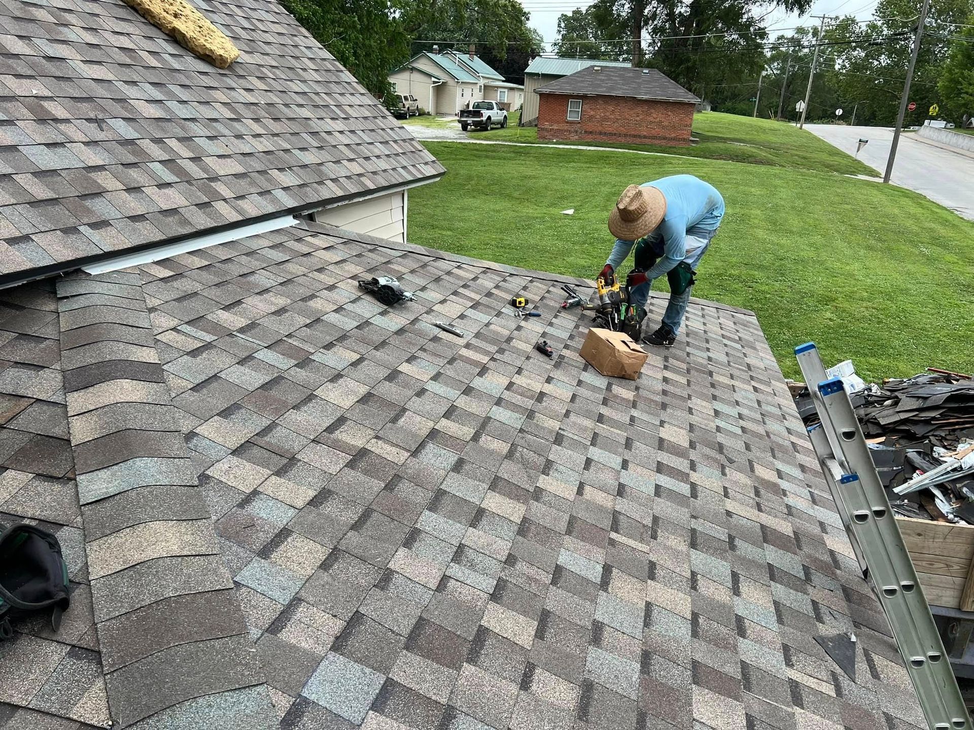 Worker kneels on a shingled roof, using tools to repair shingles beside a roadside yard.