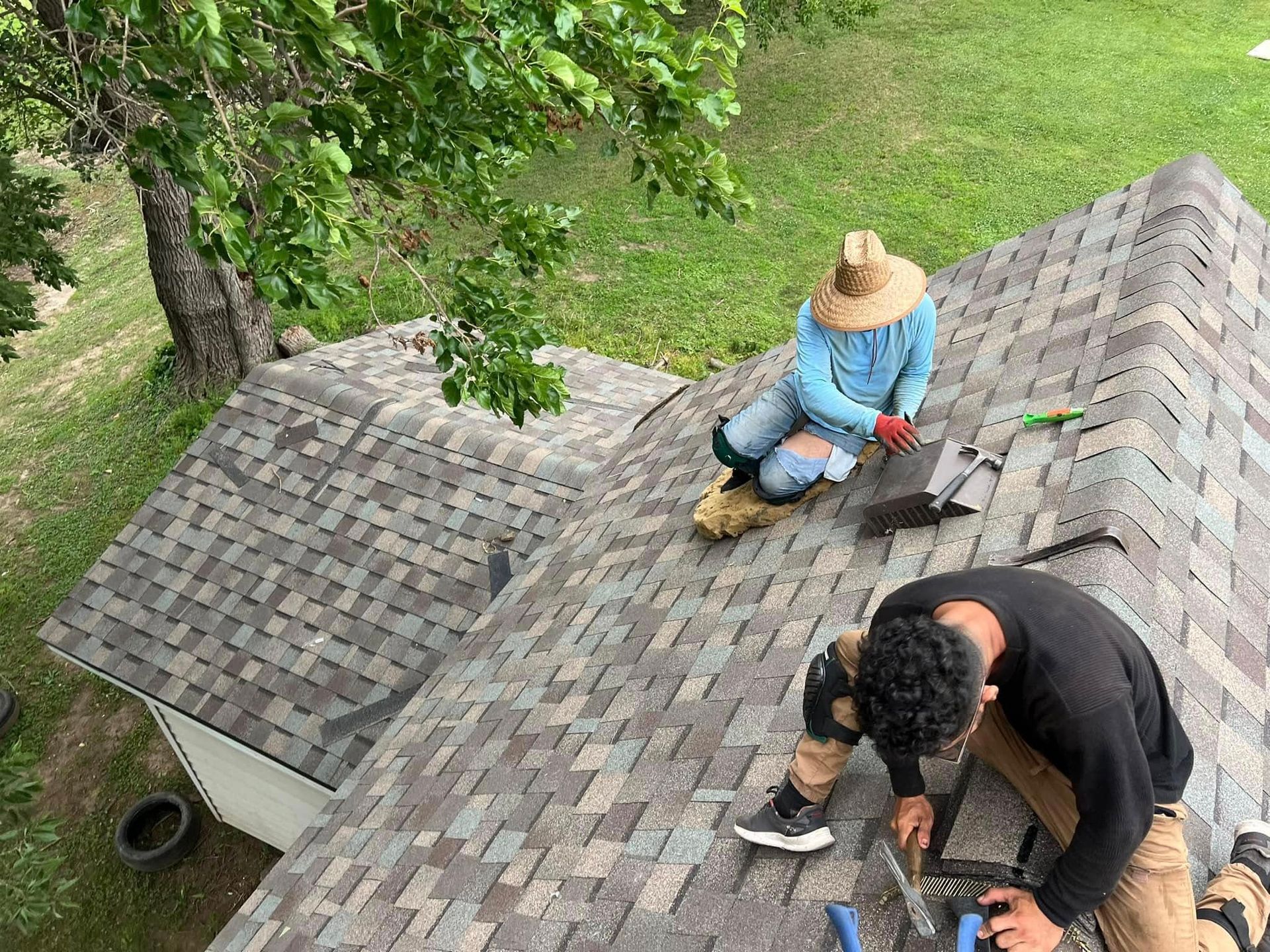 Two people kneeling on a shingled roof, inspecting or repairing it near trees and grass below.