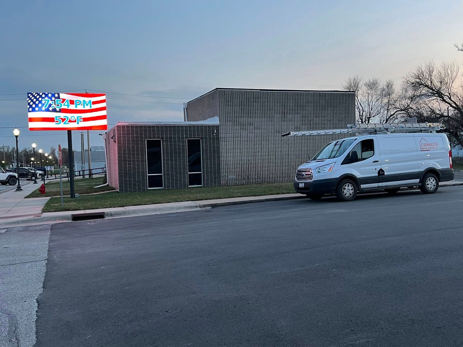 White van parked outside a small building beside an American flag sign at dusk.