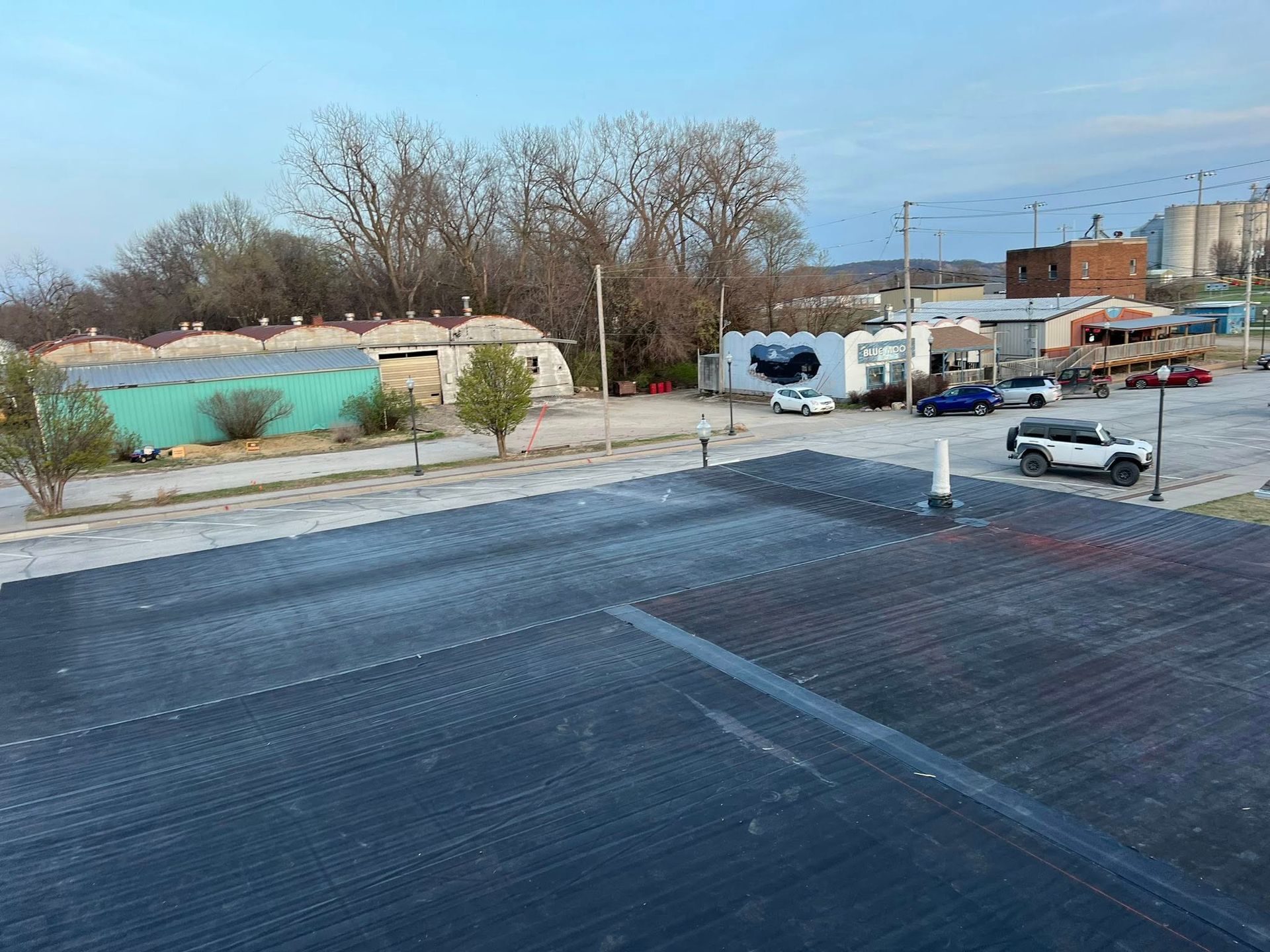Empty paved lot beside a turquoise pond, with leafless trees, parked cars, and low buildings in the distance