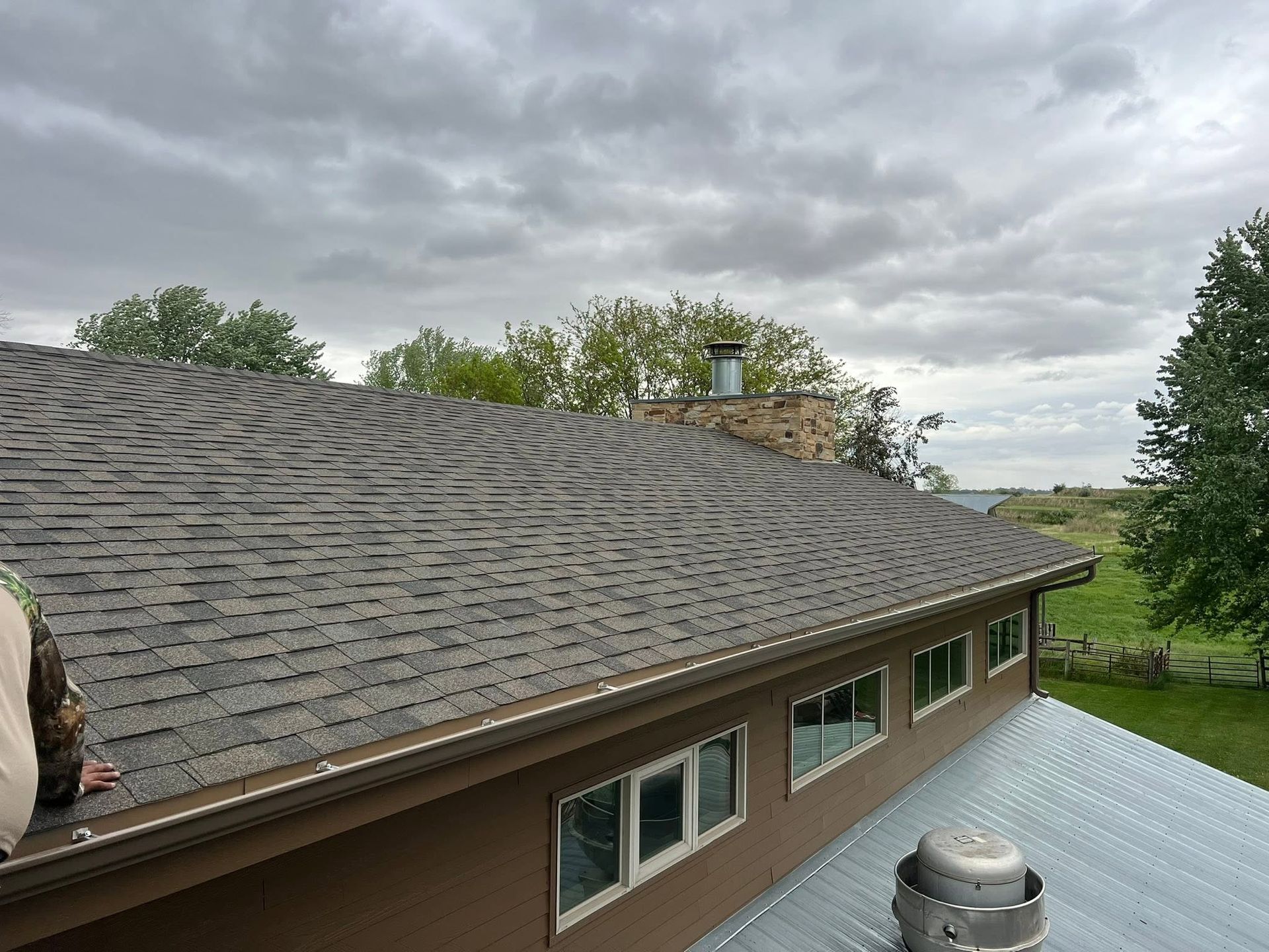 Roof of a brown house with gray shingles, a metal vent, and cloudy sky overhead