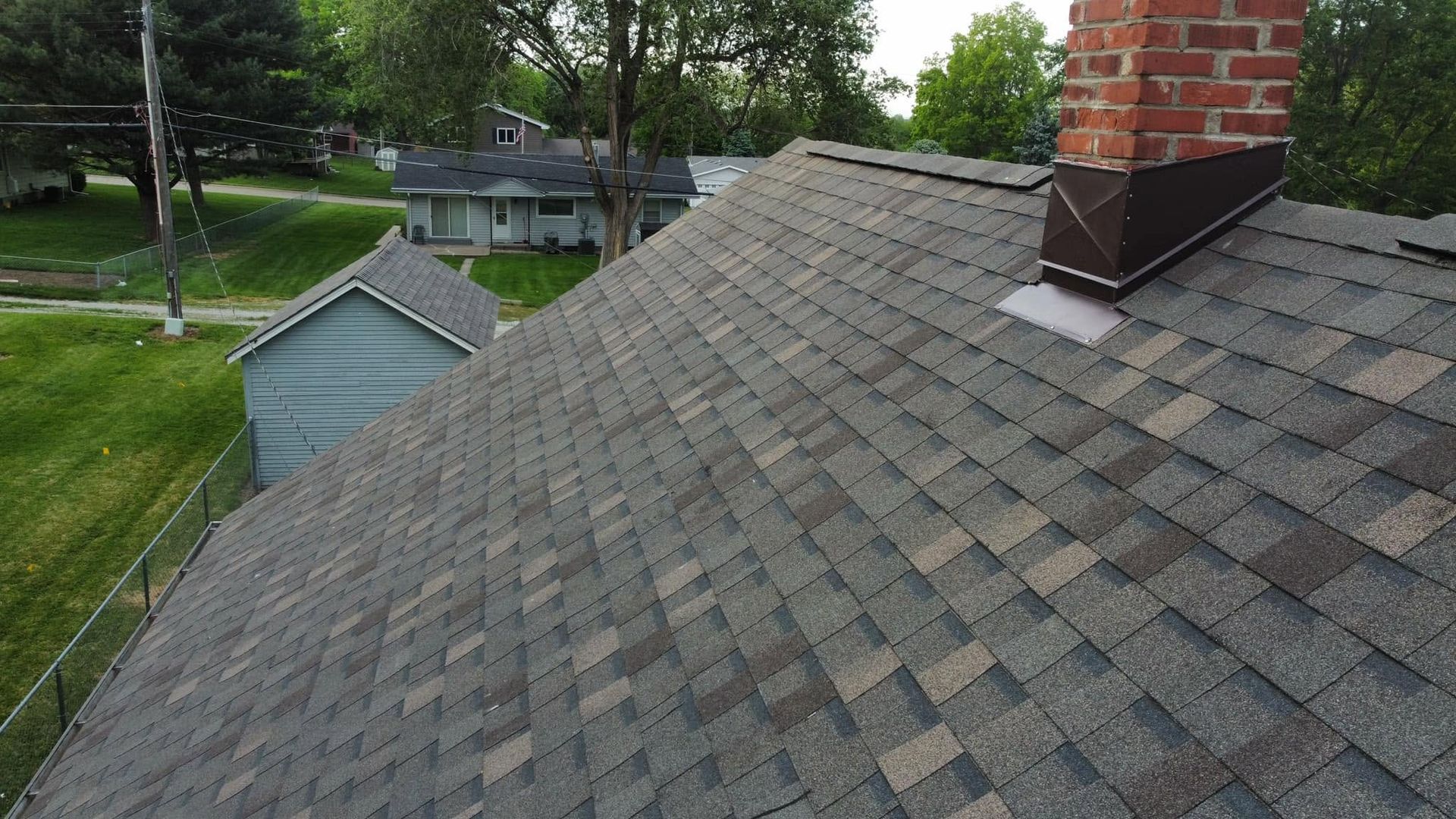 Residential shingle roof with a brick chimney, viewed from above near a green shed and trees