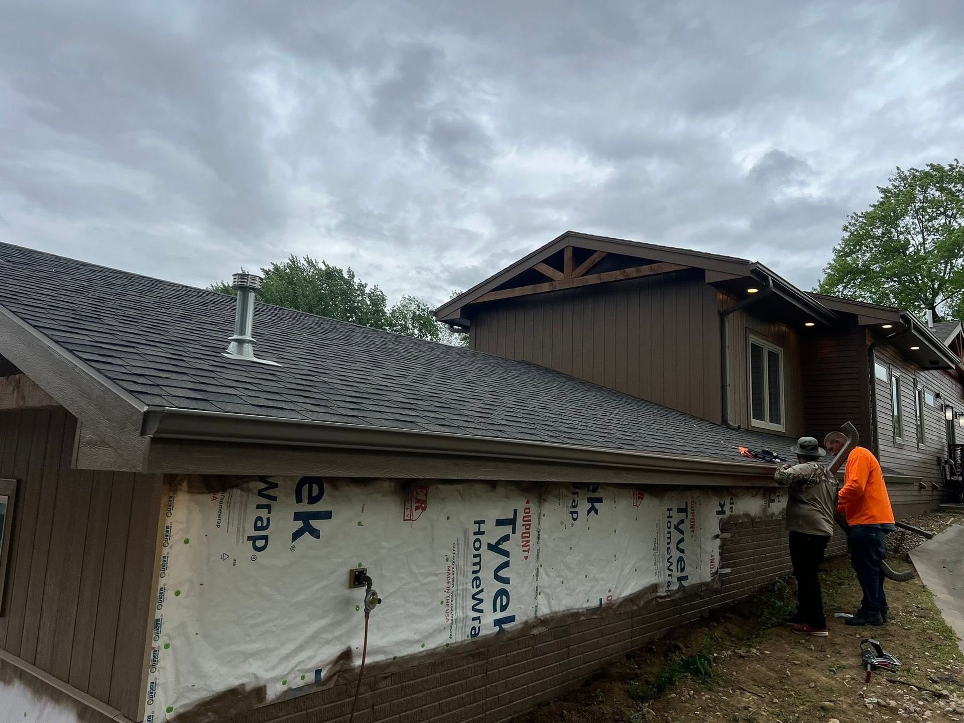 Worker in orange jacket installs siding on a house under construction beside a shingled roof.