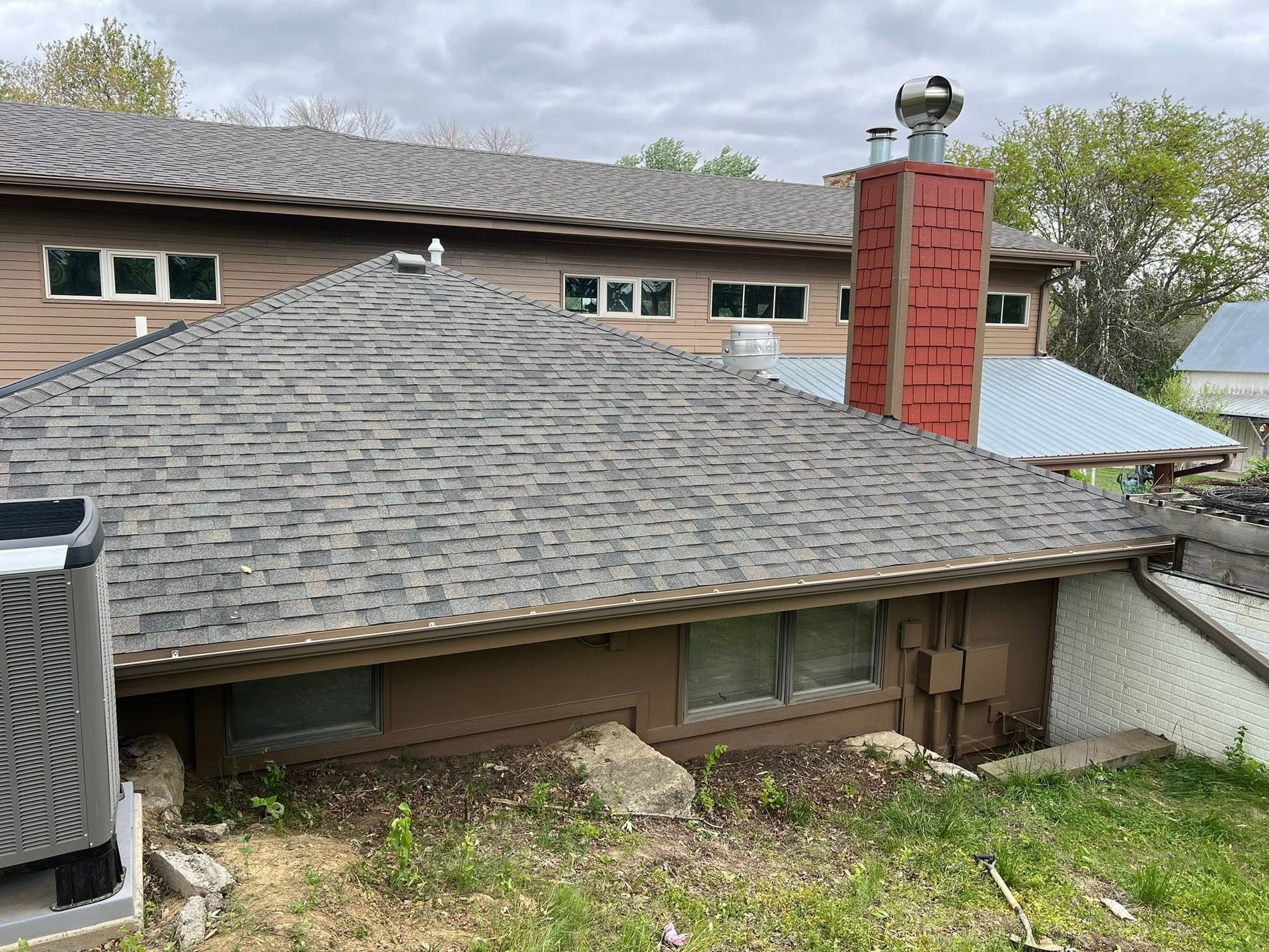 Residential rooftop with gray shingles, red brick chimney, and overgrown backyard under cloudy sky