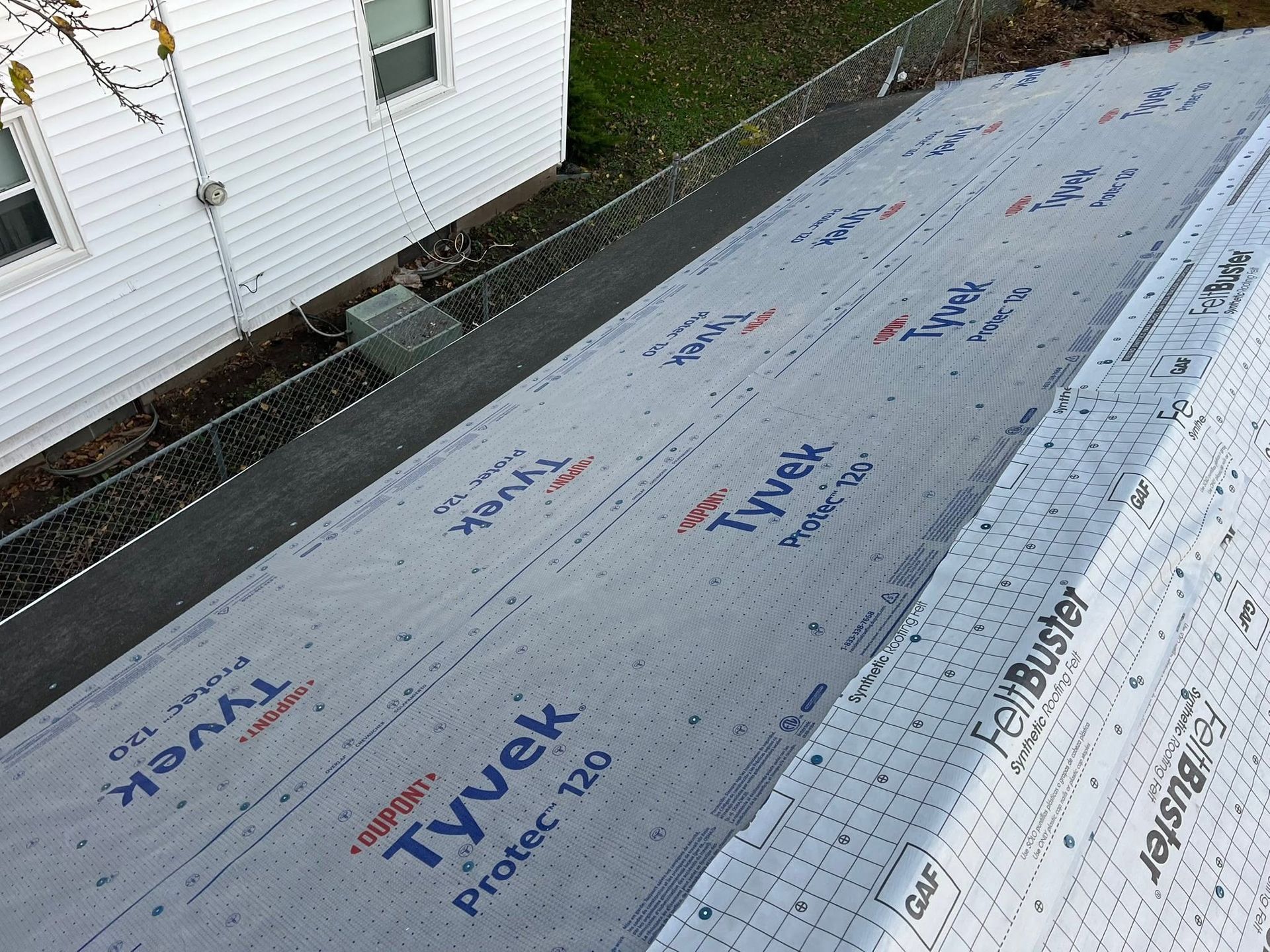 Roof underlayment being installed on a house roof beside white siding and a window.
