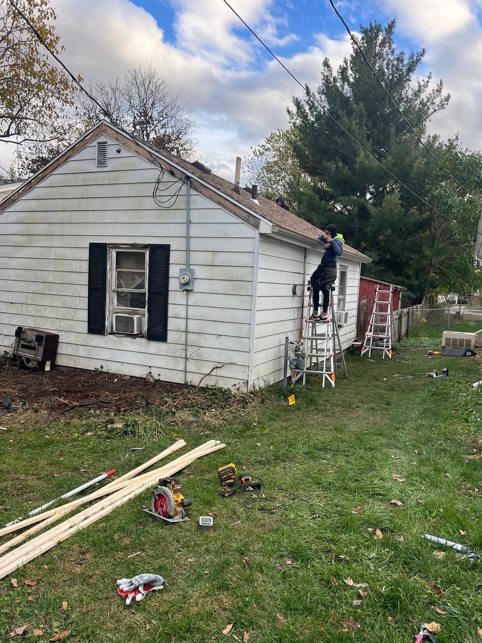 Person working on the exterior of a small white house with siding and debris in the yard