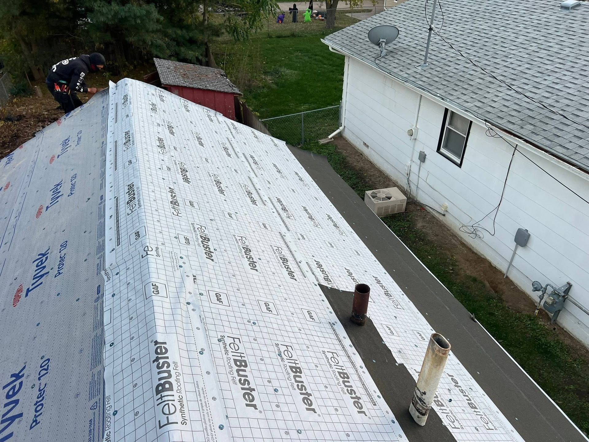 Roof covered with white underlayment next to a white house and yard, with vents and a chimney visible.