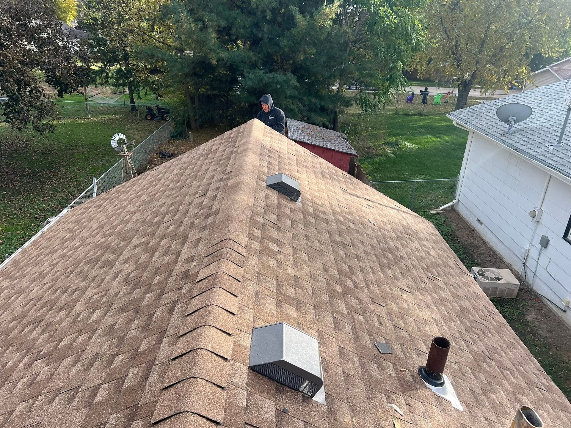 View from rooftop with tan shingles, vents, and a person at the ridge, overlooking trees and nearby houses