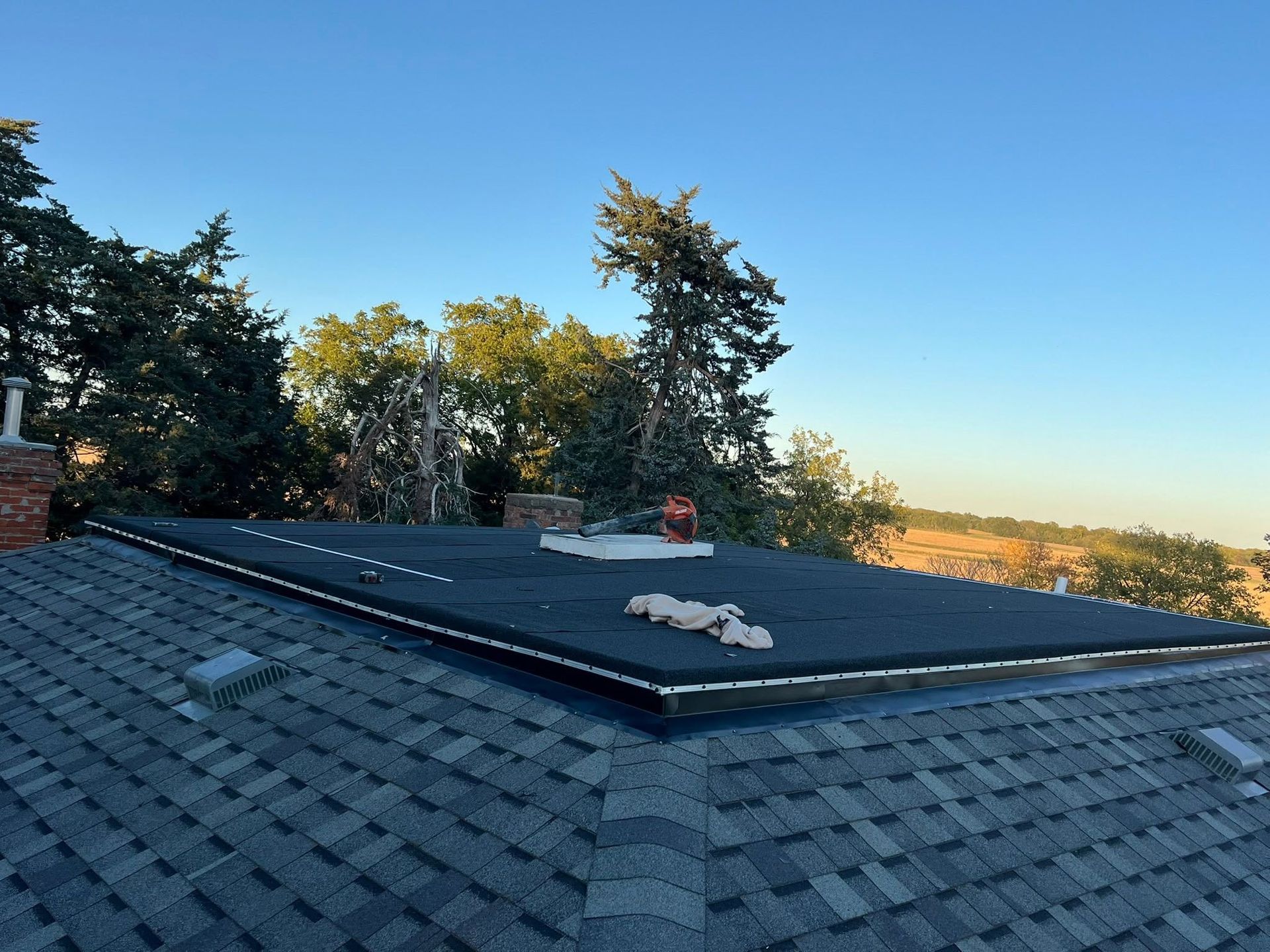 Rooftop shingles with a flat vent cover, framed by trees under a clear blue sky.