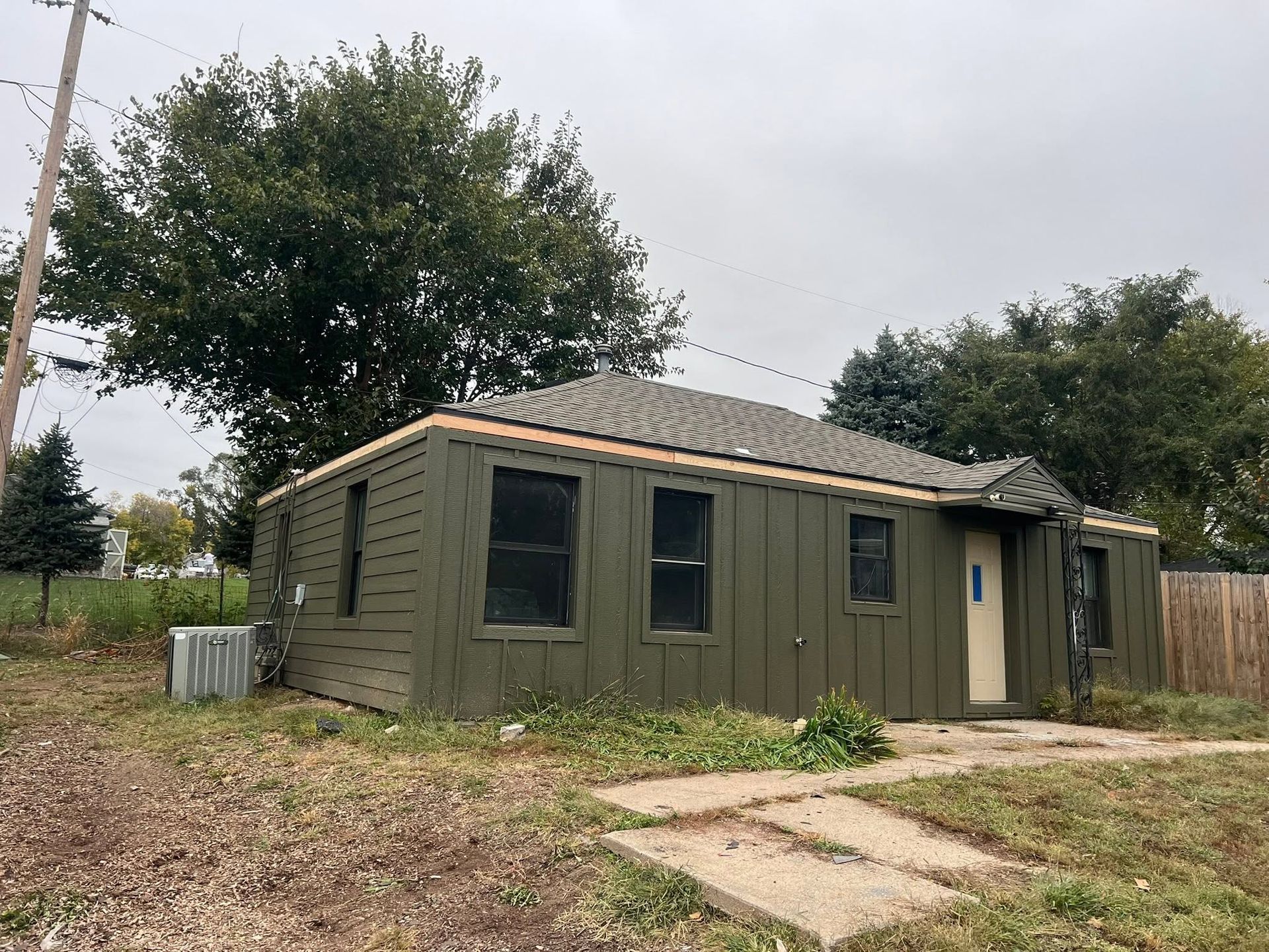 Small olive-green outbuilding with windows and a side door in a grassy yard under cloudy skies