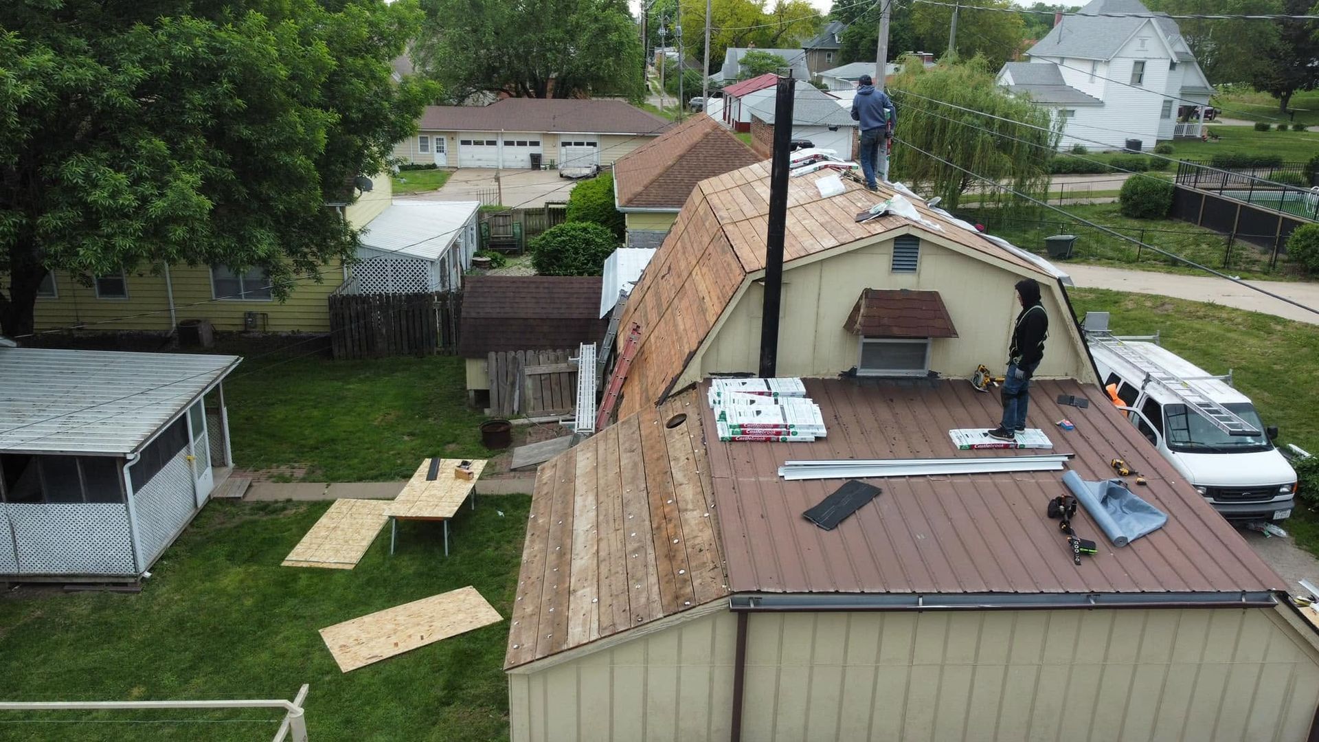 Rooftop under repair with exposed plywood, vents, and a ladder beside a fenced backyard and neighboring houses
