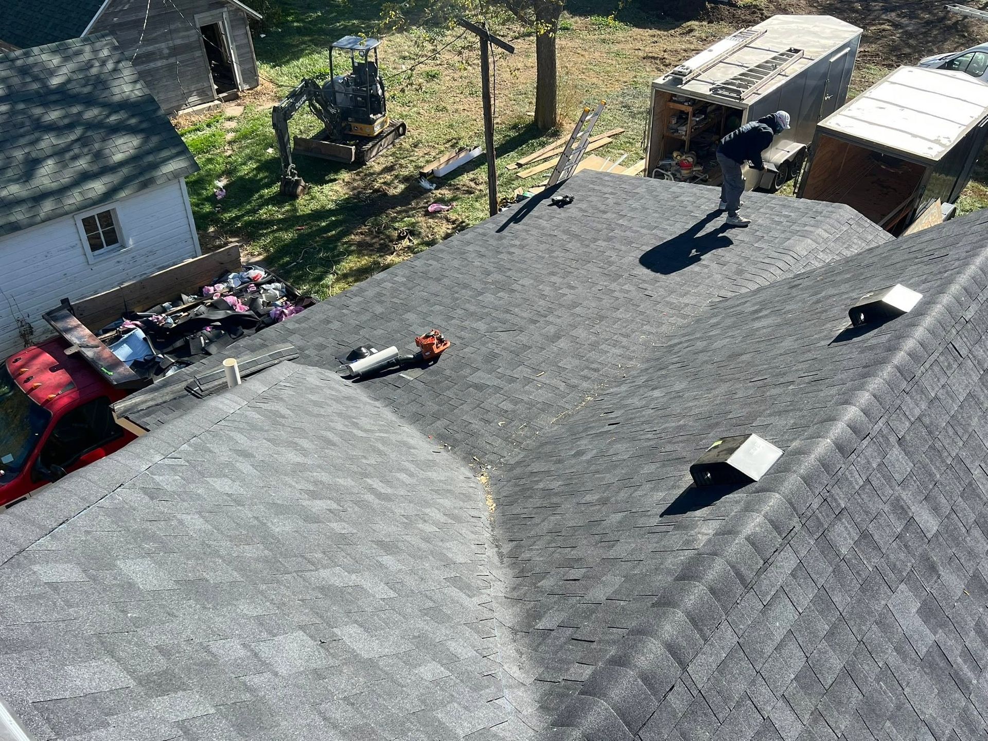 Aerial view of a gray shingled roof with roof vents, surrounded by trees and a yard with vehicles and equipment.