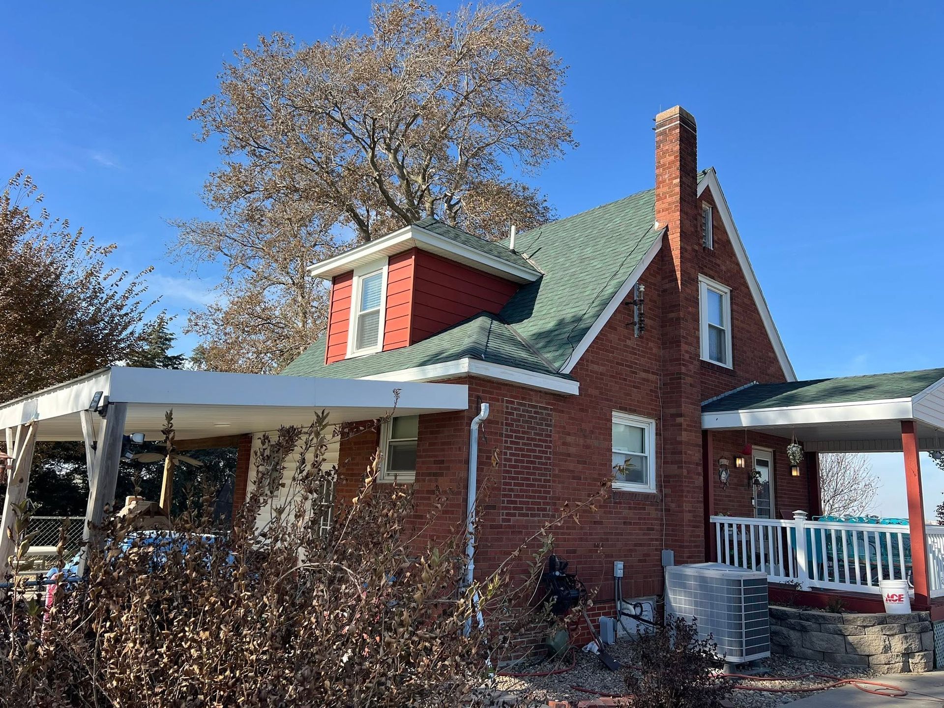 Brick house with green roof, white porch, and leafless trees under a clear blue sky