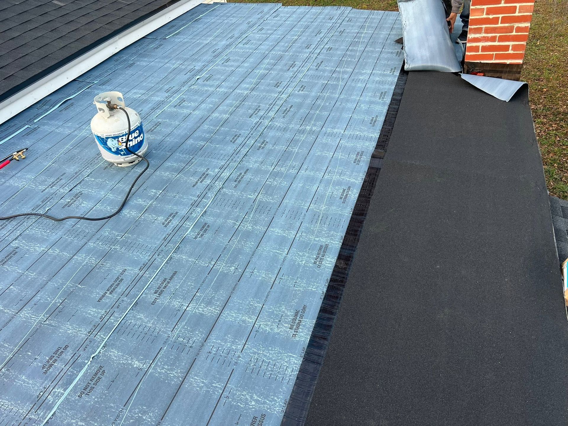 Rooftop with blue membrane roofing, black underlayment strip, and a gas canister near a brick chimney.