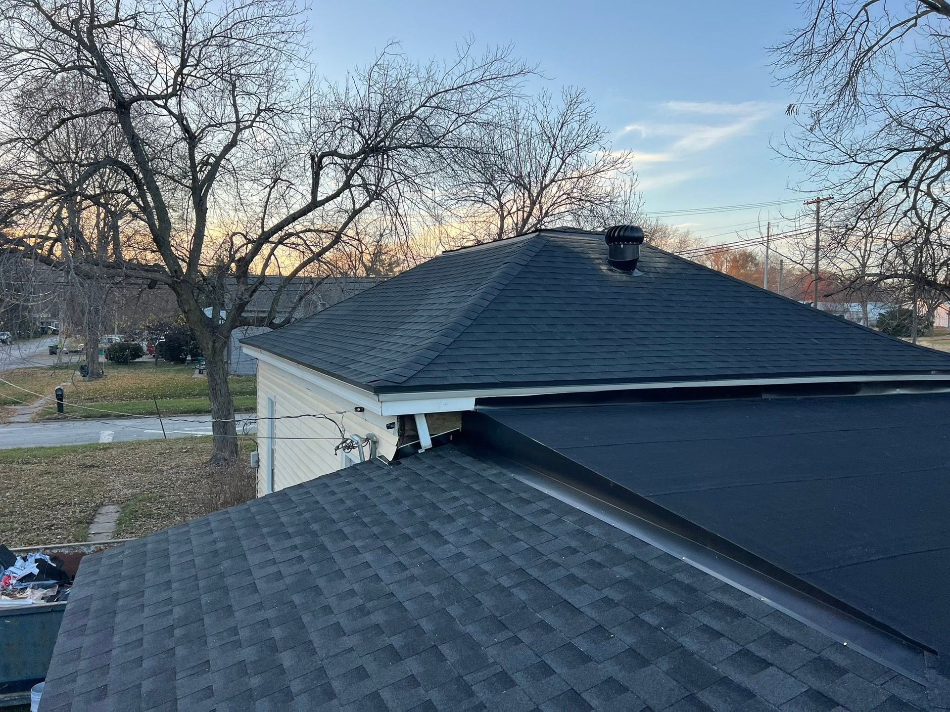 House roof being repaired with dark shingles at sunset, with bare trees in the background