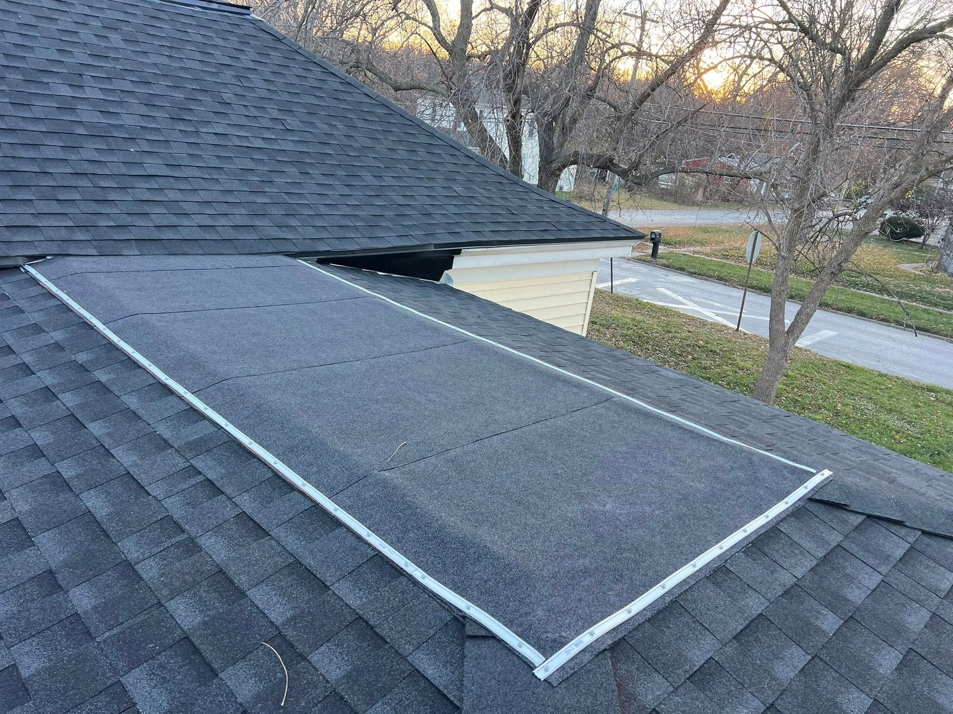 Roof skylight with a raised rectangular cover on a shingled house roof, near trees and a road.