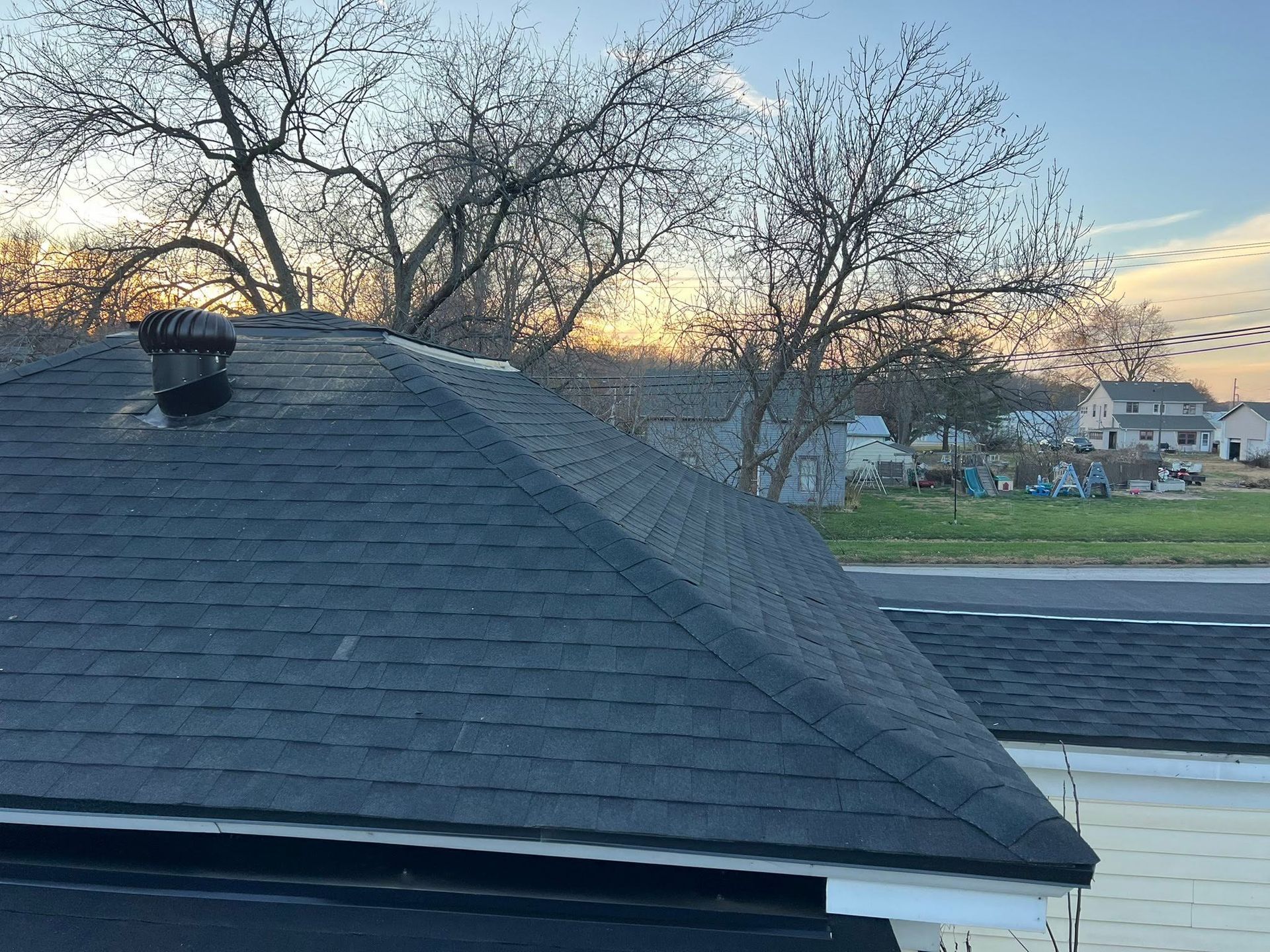 Rooftop with dark shingles at sunset, leafless trees, and neighboring houses in the background.