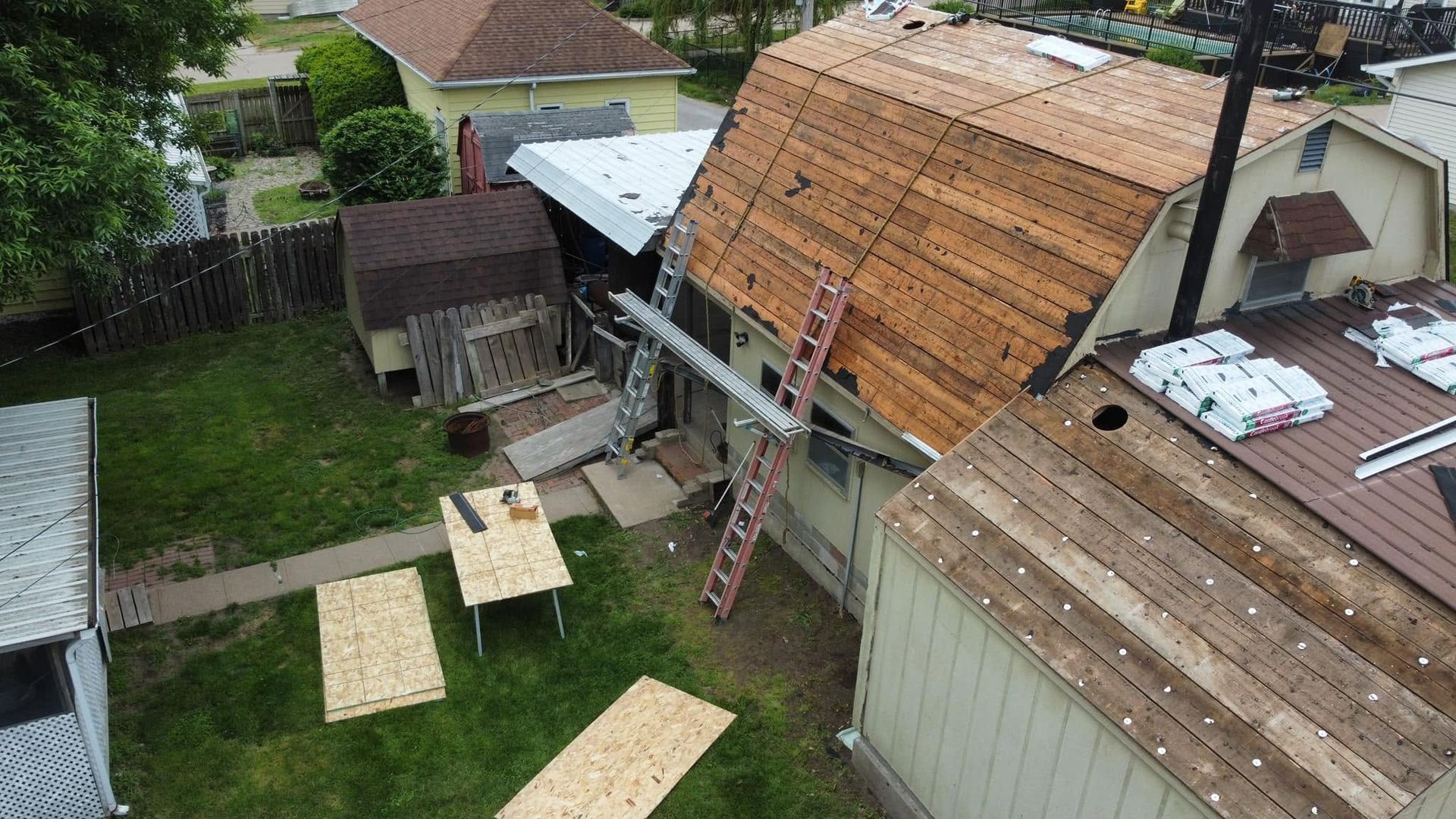 Backyard with a steep roof under repair, ladder, planks, and a small work area between sheds and a house.