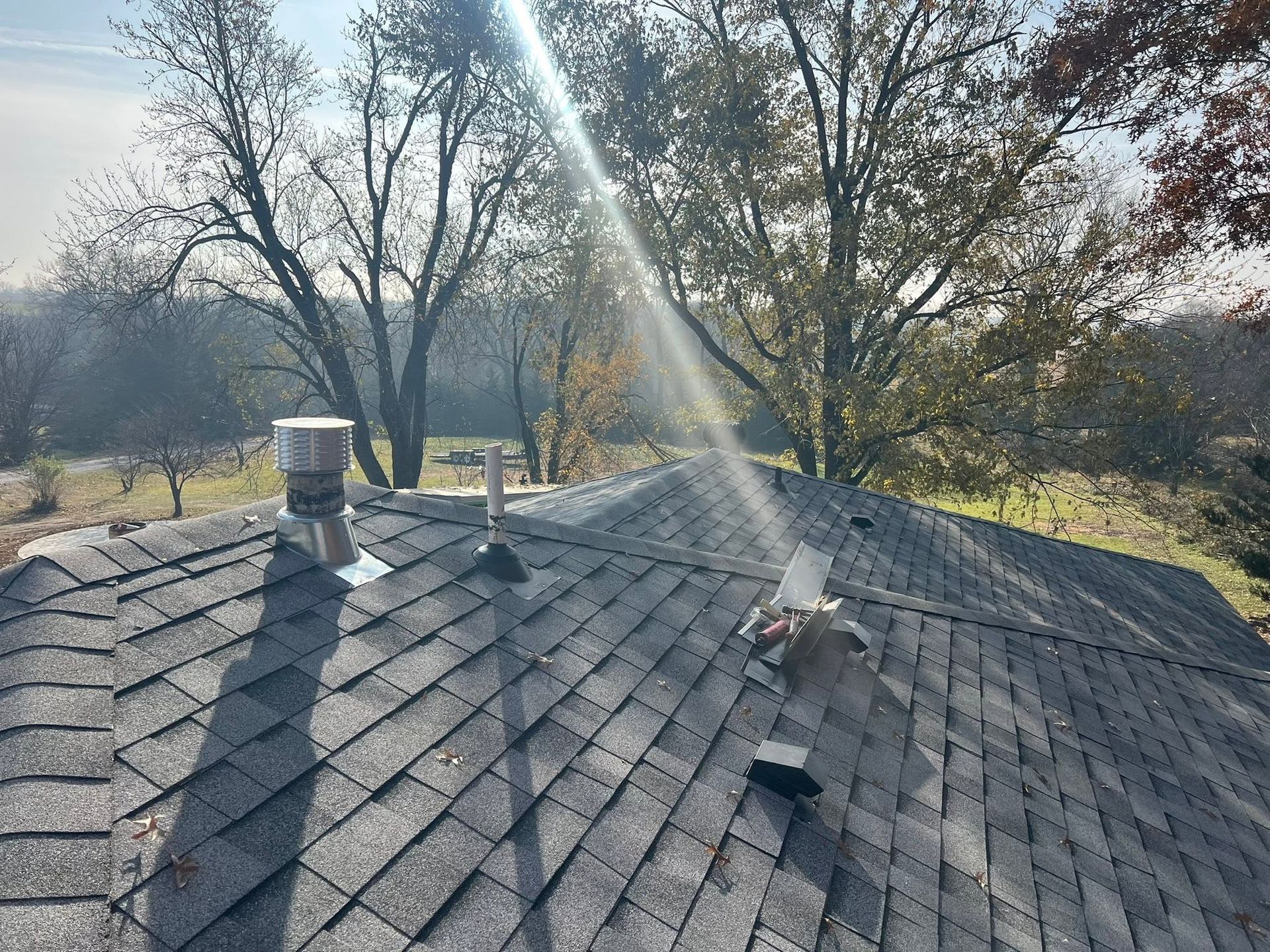 Sunlit shingled roof with vents, trees in the background, and a bright diagonal sunbeam