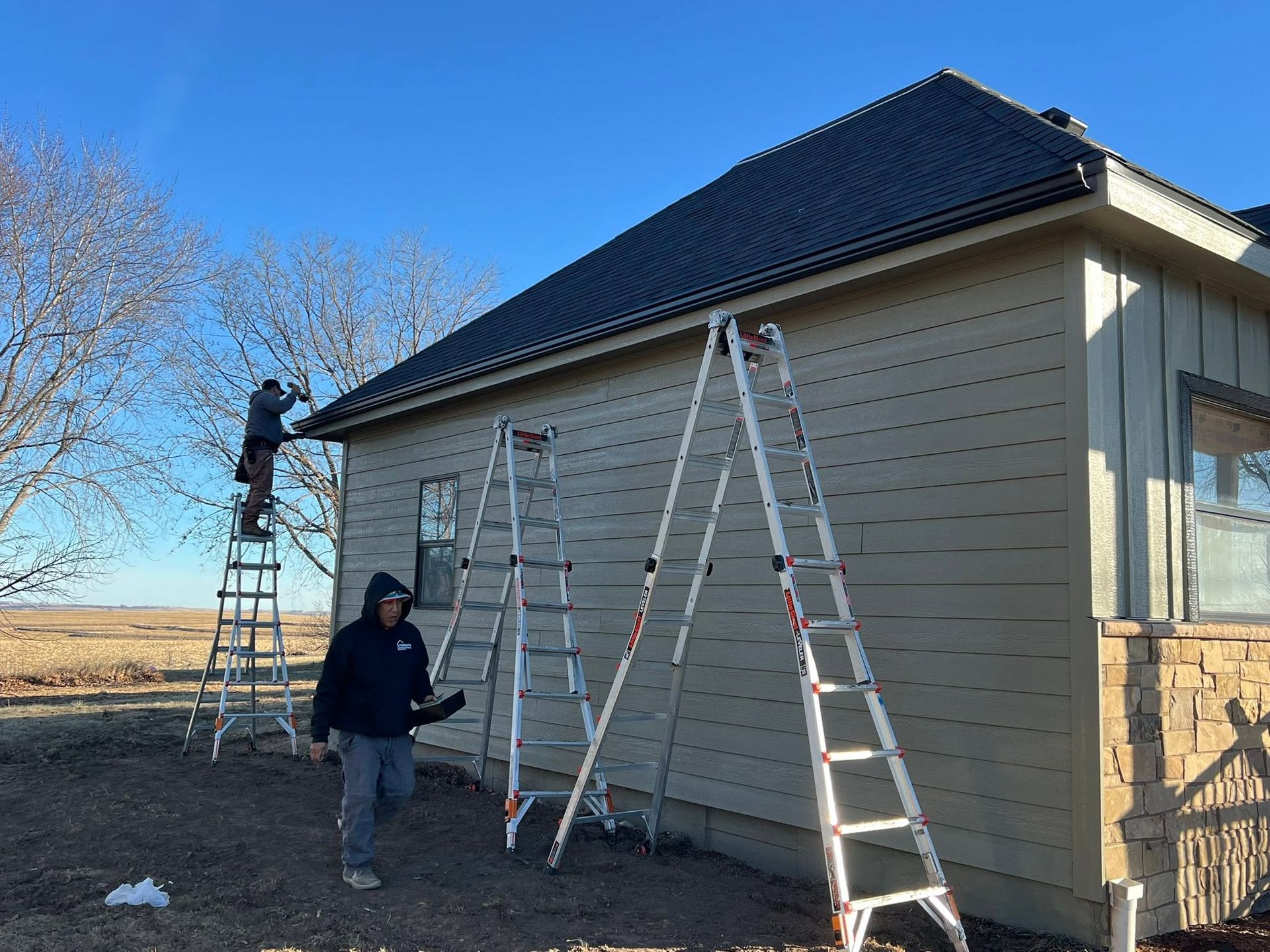 Workers on ladders painting the side of a beige house with a dark roof outdoors.