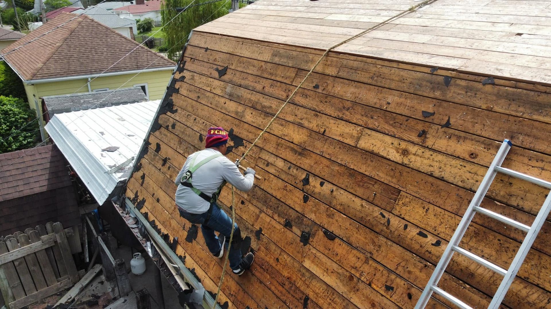 Worker installing shingles on a steep roof beside a ladder and nearby houses