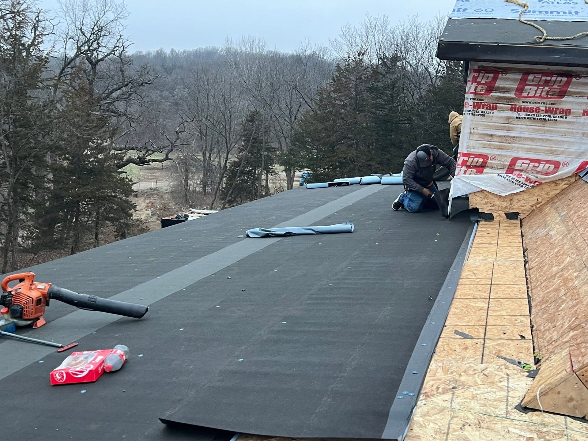 Roofing work in progress on a house, with black underlayment, tools, and a worker near the roof edge.