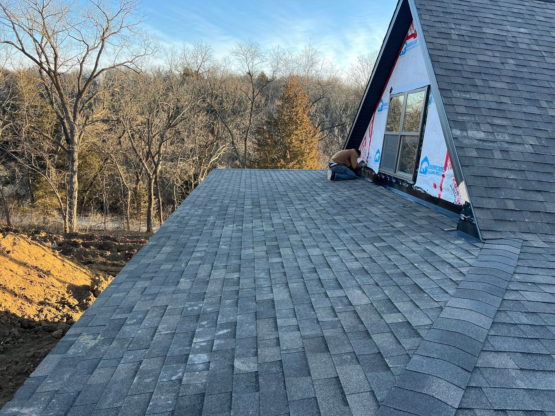 Roof under construction with gray shingles, worker near an A-frame dormer, and leafless trees in the background