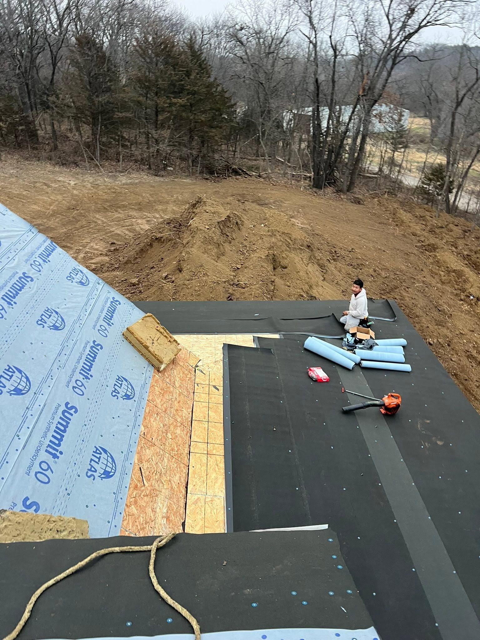 Roof under construction with plywood, black underlayment, blue insulation, and tools; worker sitting near materials