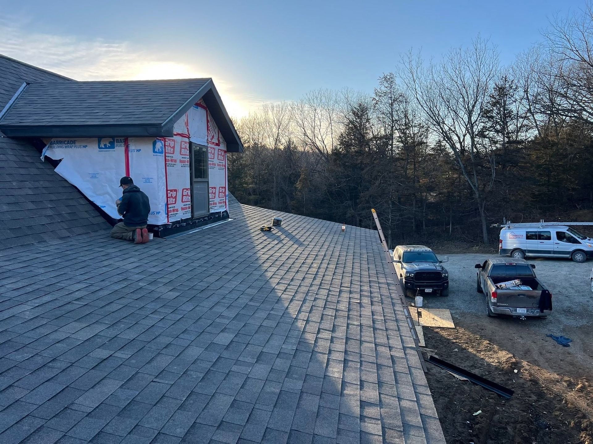 Roof under construction with exposed sheathing and workers beside parked vehicles at dusk