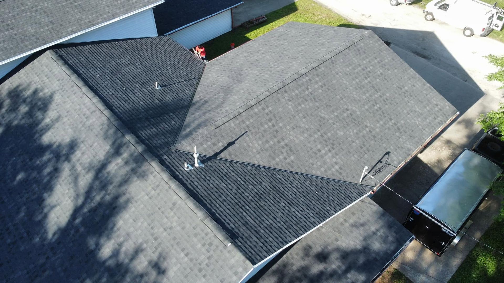 Aerial view of a gray shingled house roof with vents and skylights, beside a driveway and parked vehicle.