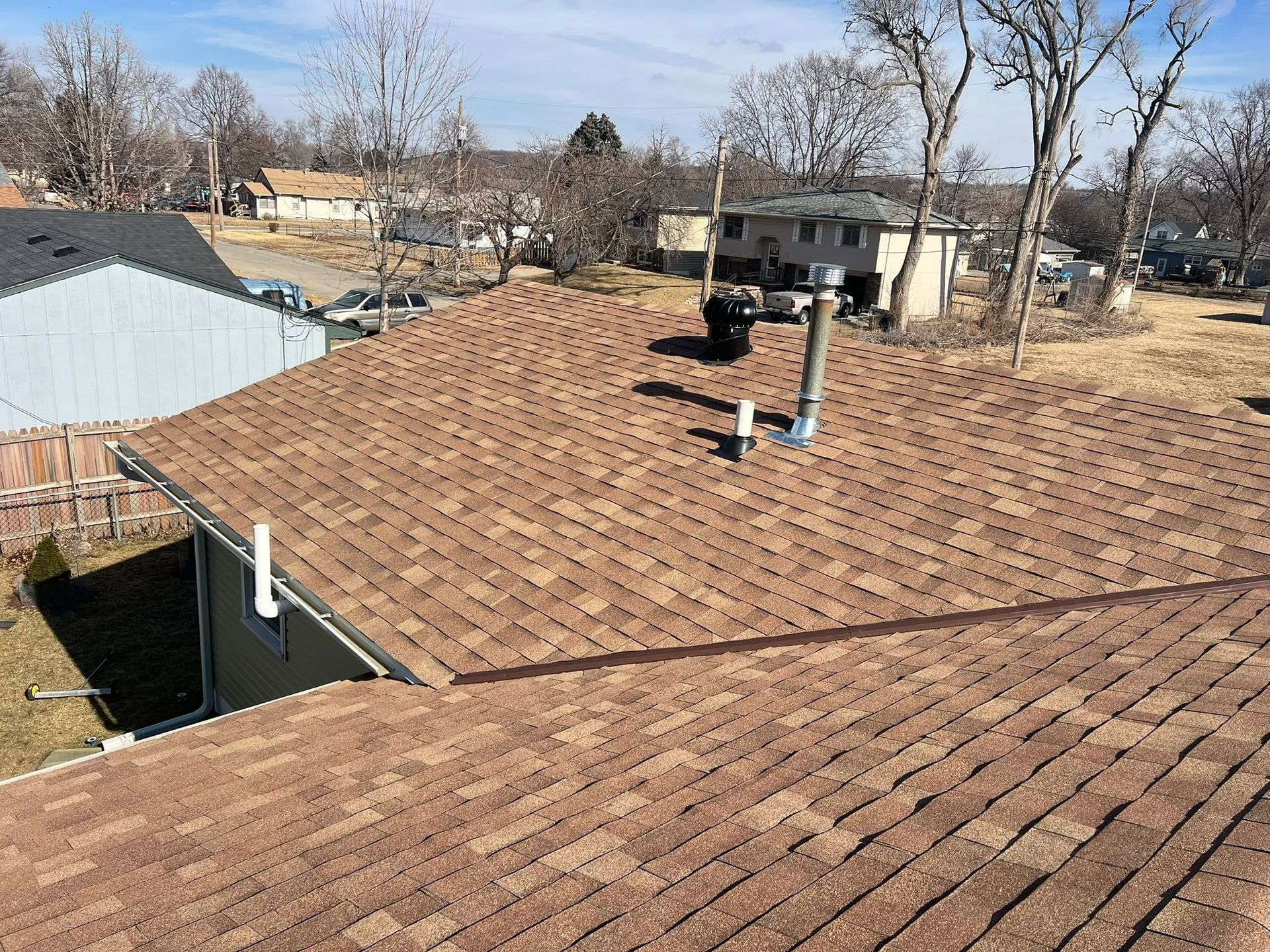 Brown shingle roof with two vent pipes in a residential neighborhood under clear sky