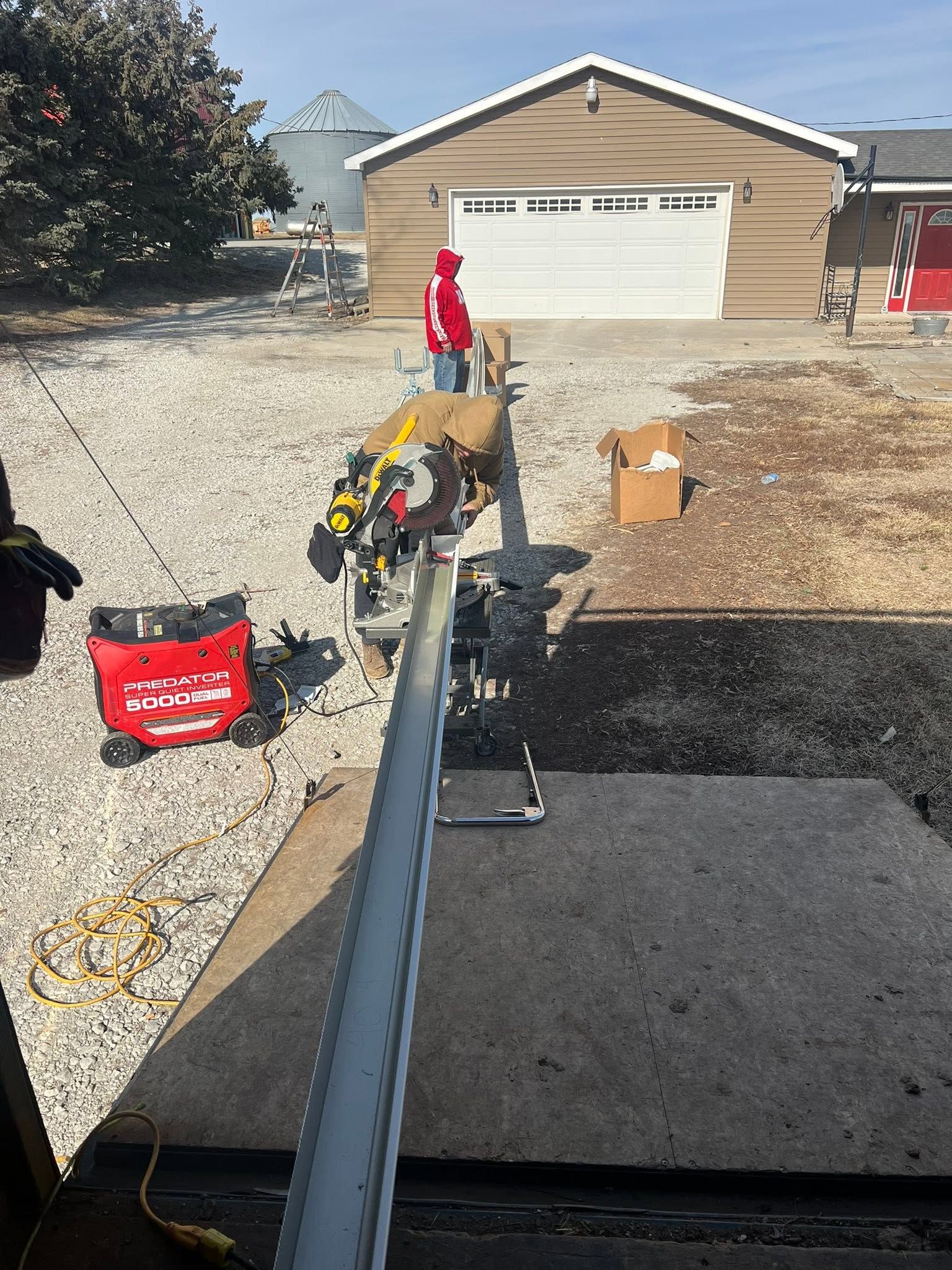 Person in red jacket using a long pole on a gravel driveway near a garage, with tools scattered nearby.