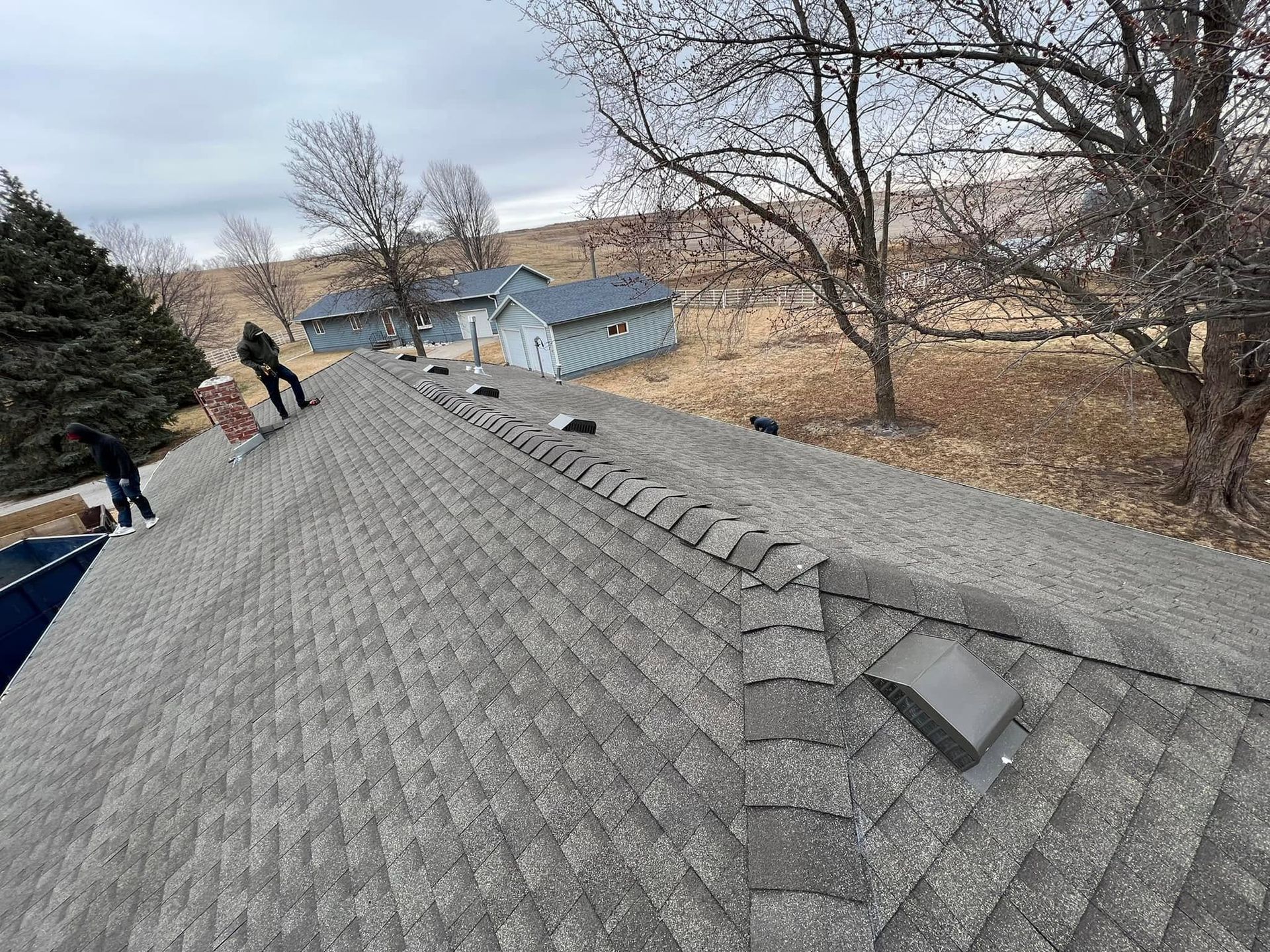 Roofing workers on a shingled roof, with trees and a house in the background.