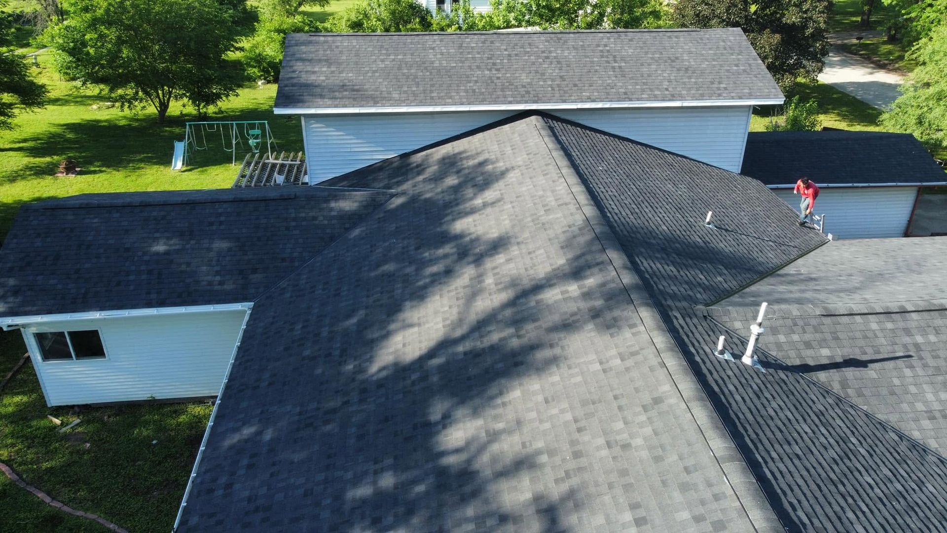 Aerial view of a gray shingled roof with a person in red standing near the edge by a driveway and garage.