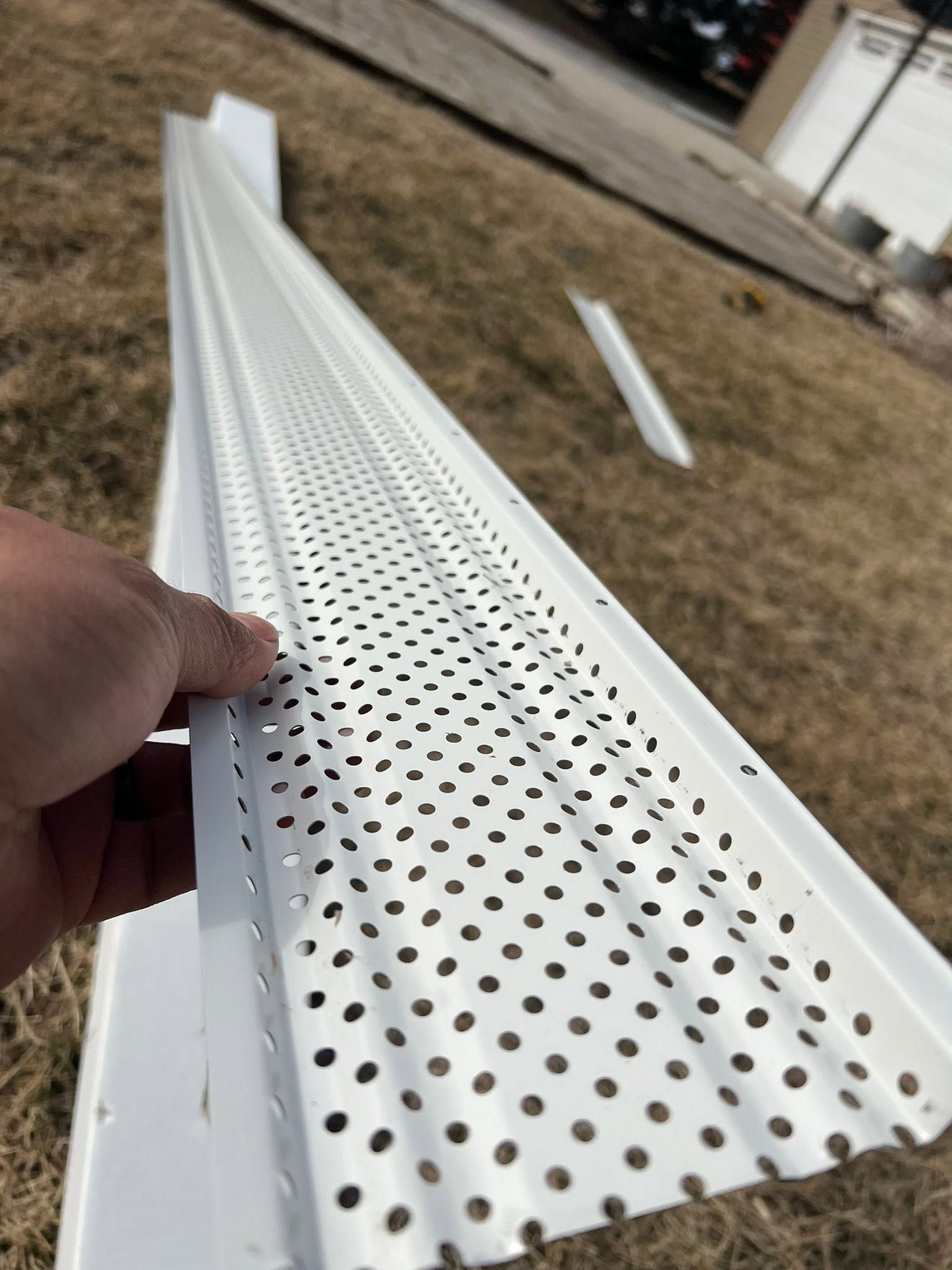 Hand holding a white perforated metal gutter or trim piece outdoors on grass