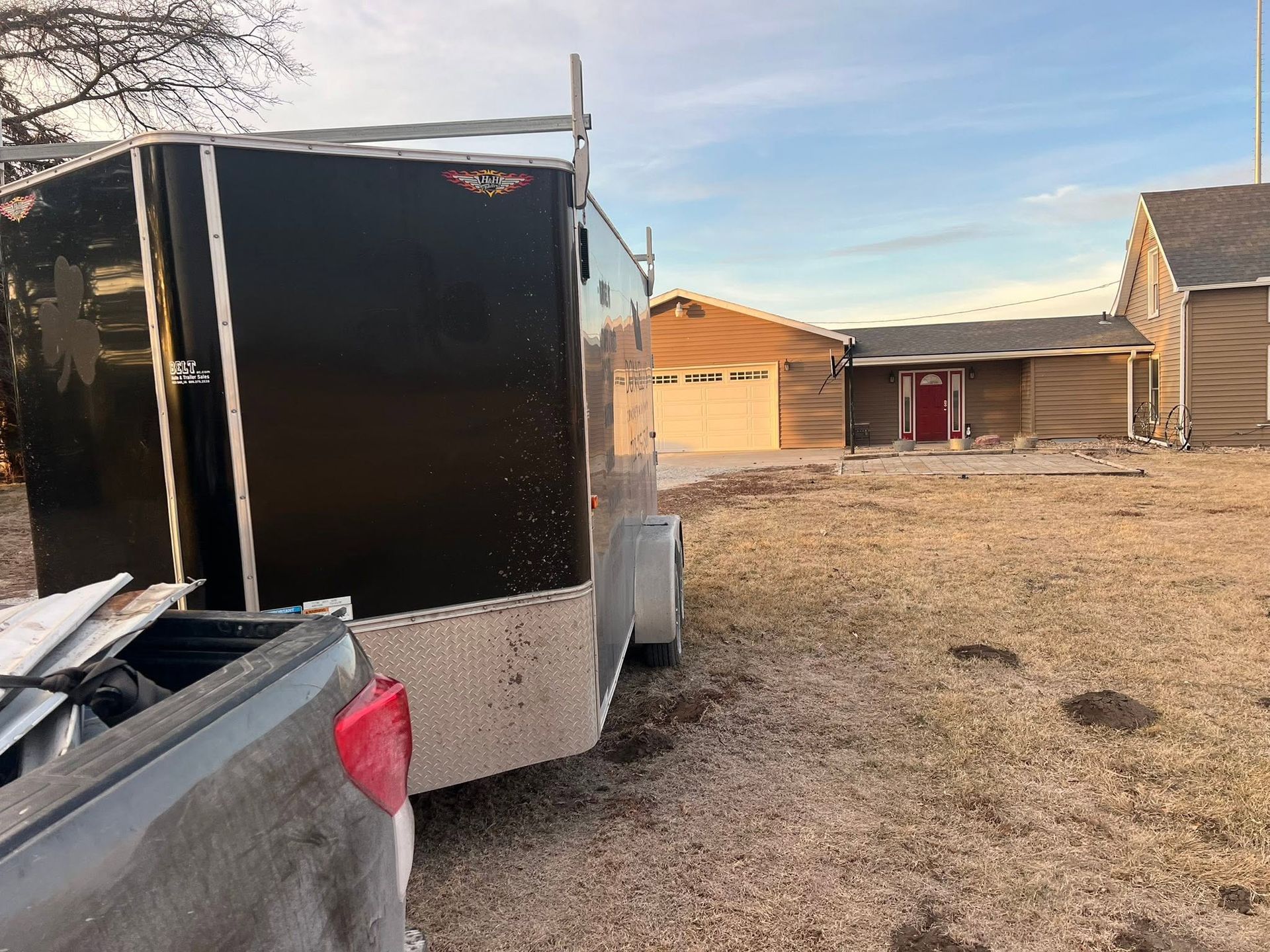 Black utility trailer beside a pickup truck in a dirt yard with houses in the background